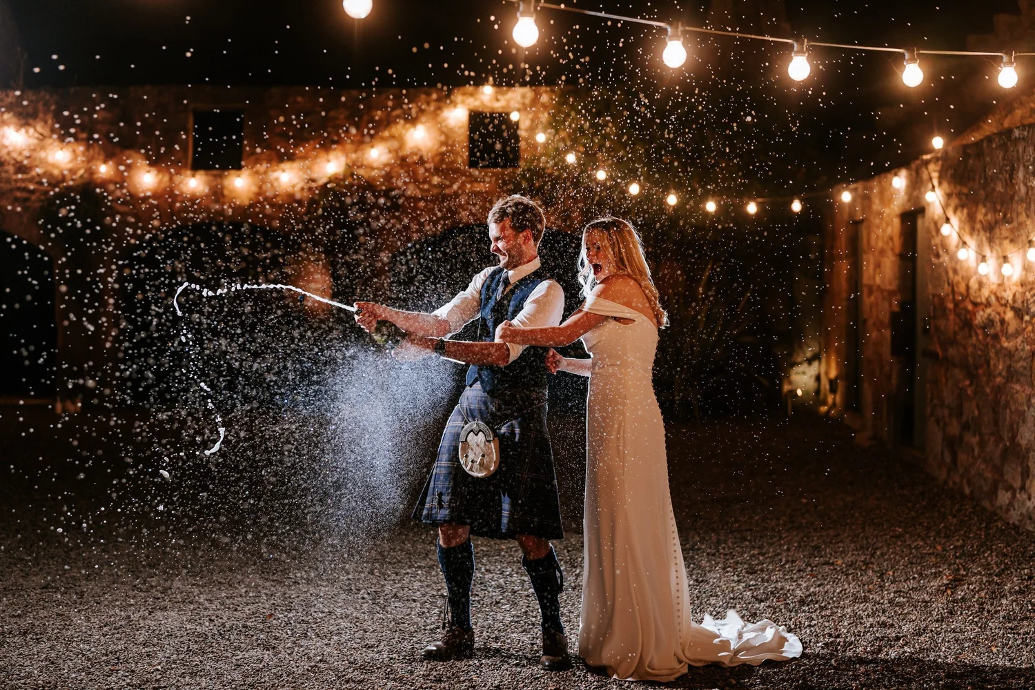 The couple spray a bottle together beneath the courtyard lights, with droplets filling the night air.