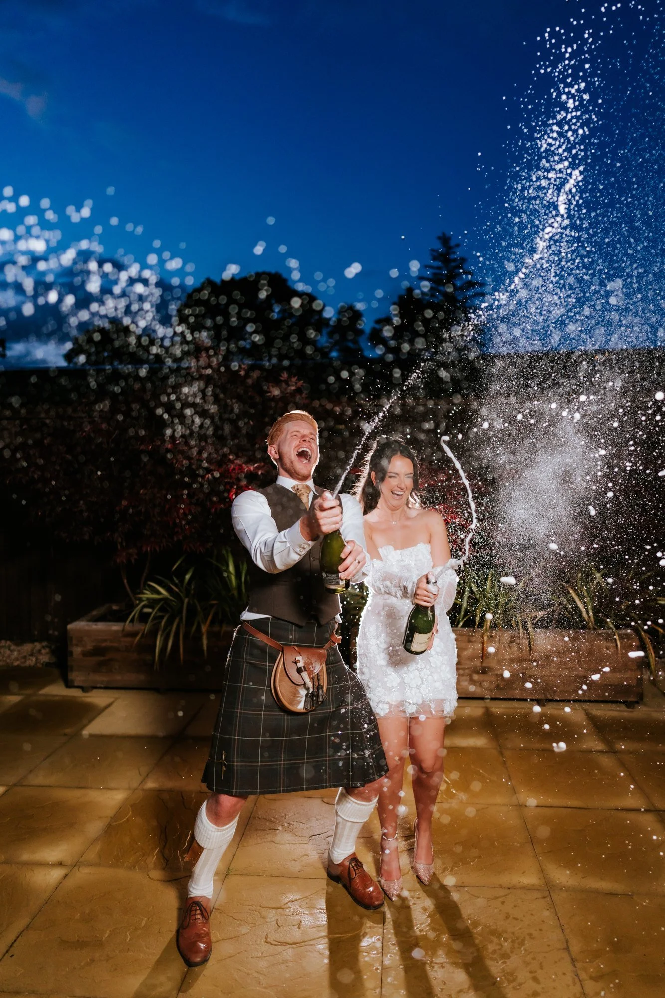 Kaitlyn and Scott laughing as they spray champagne on the terrace at dusk at Glenbervie House.