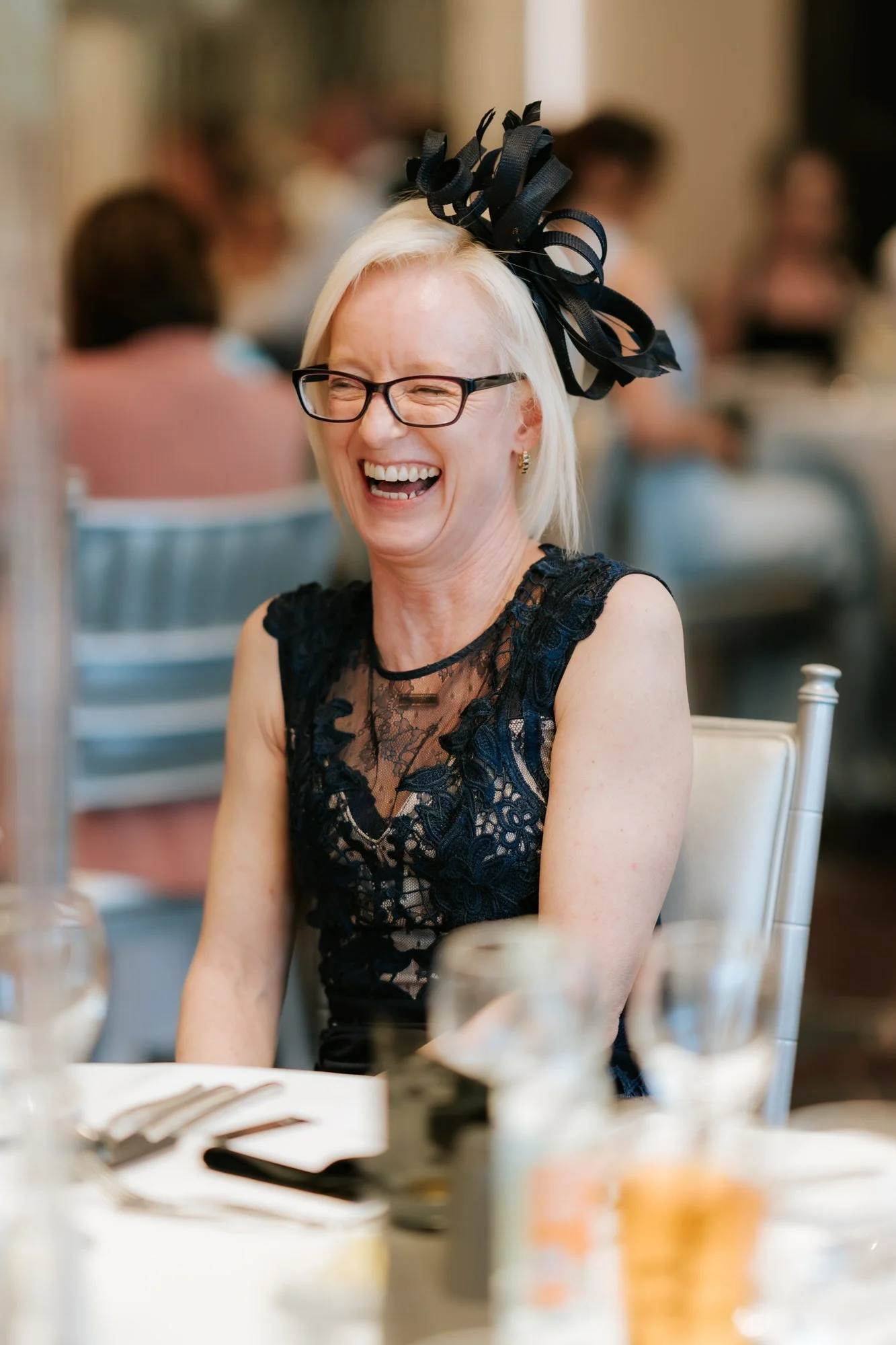A guest wearing glasses and a black fascinator laughing at the table
