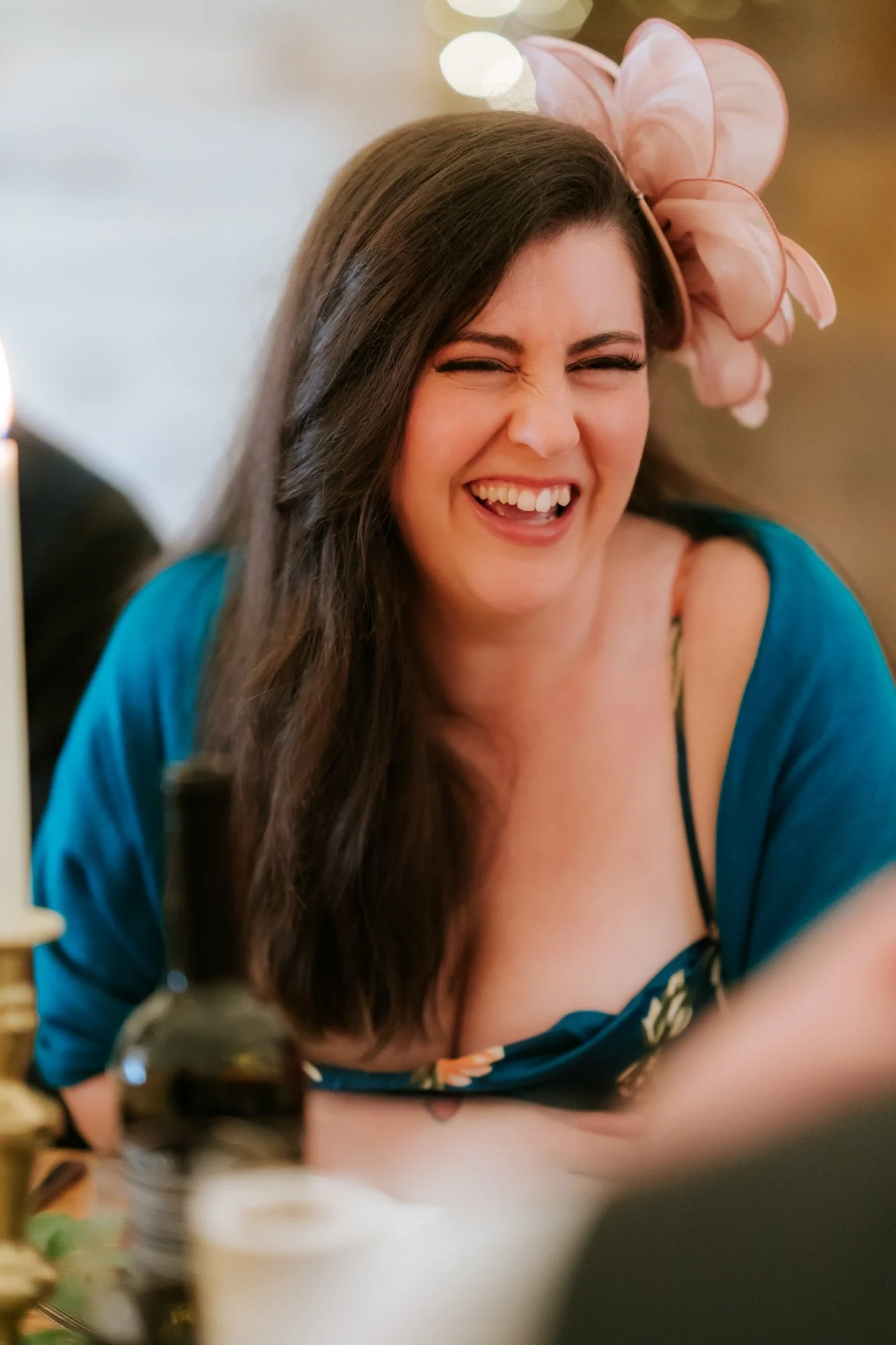 A guest in a teal outfit and pink headpiece laughs at the reception table.