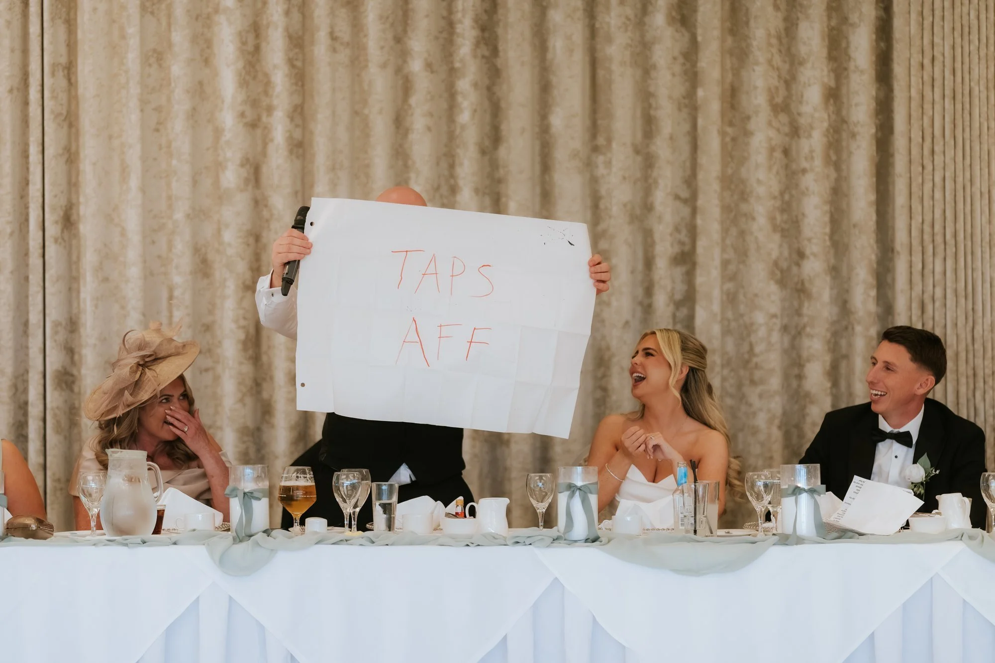 Taylor’s dad holding up his handwritten “TAPS AFF” poster as Taylor and Aiden laugh during the wedding speeches.