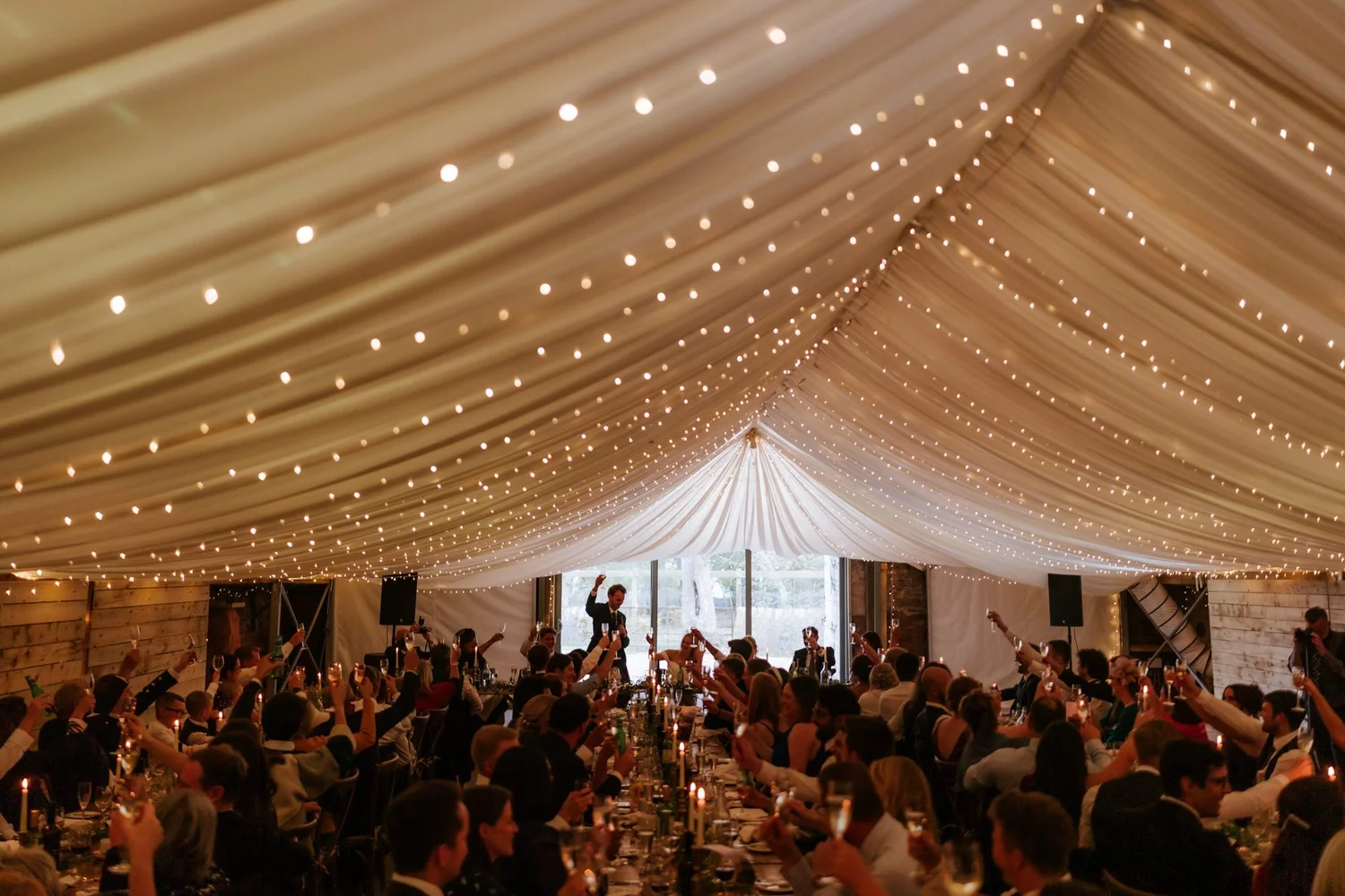 A wide view of the wedding reception, with guests raising their arms beneath a canopy of fairy lights.