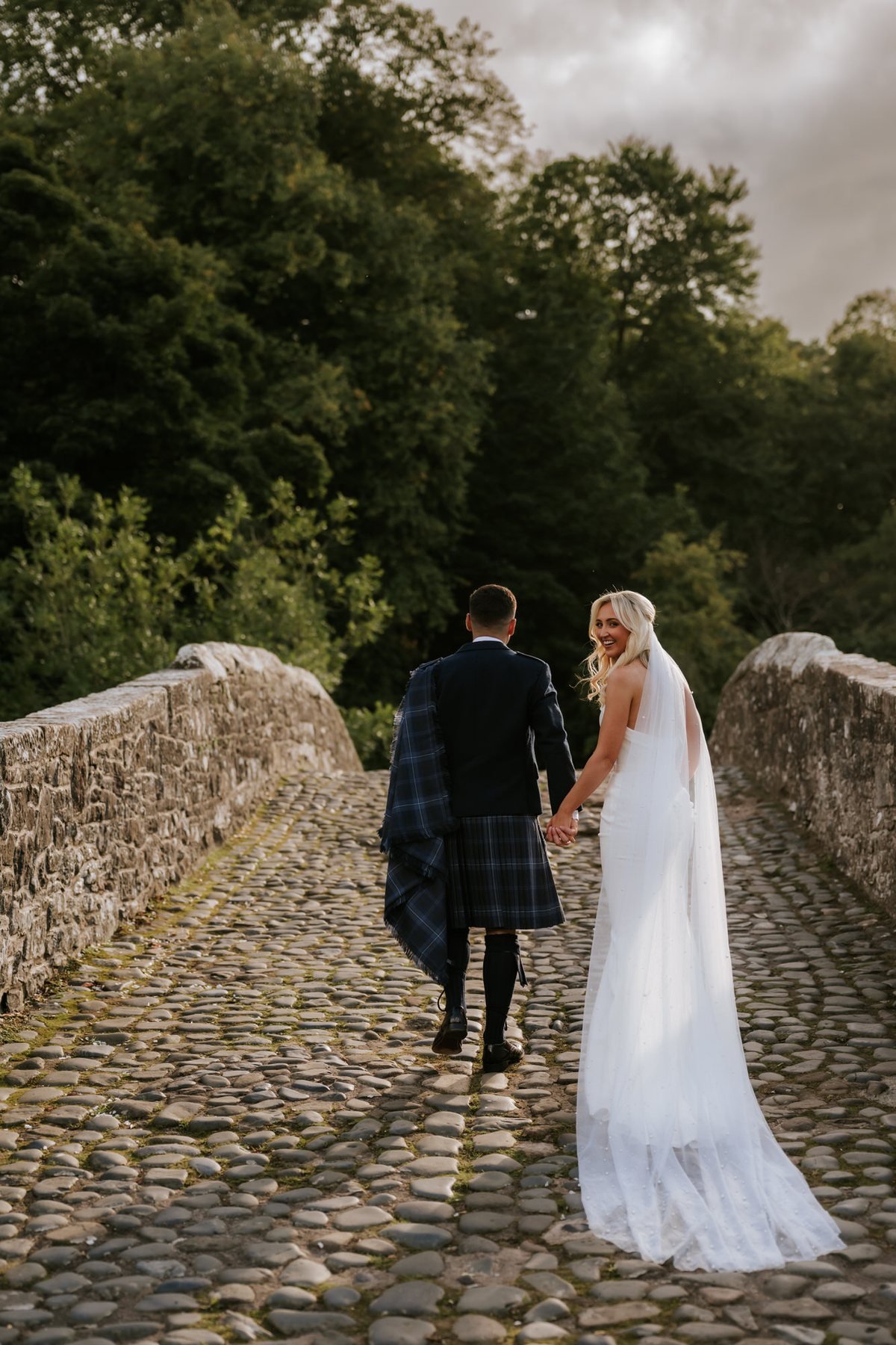 Lauren and Zen holding hands on the sunlit stone bridge in Alloway.