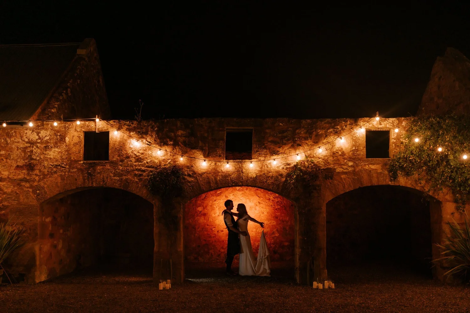 The couple face one another in the illuminated courtyard after dark, framed by stone arches and festoon lights.