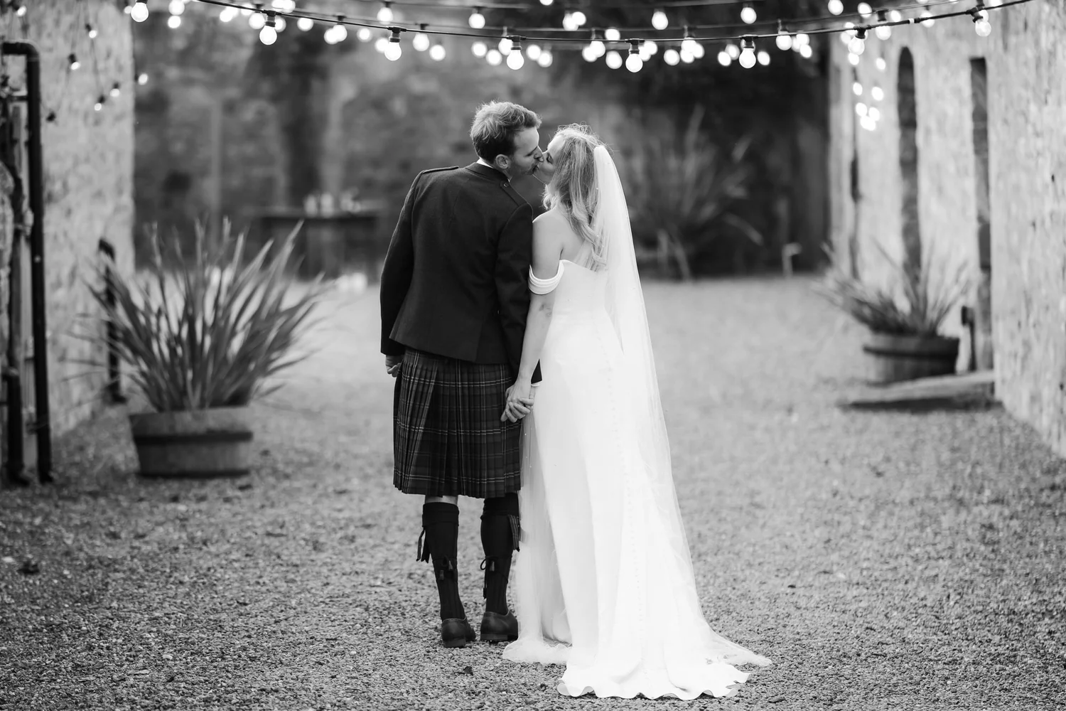 Black-and-white photograph of the couple kissing beneath festoon lights in the courtyard.