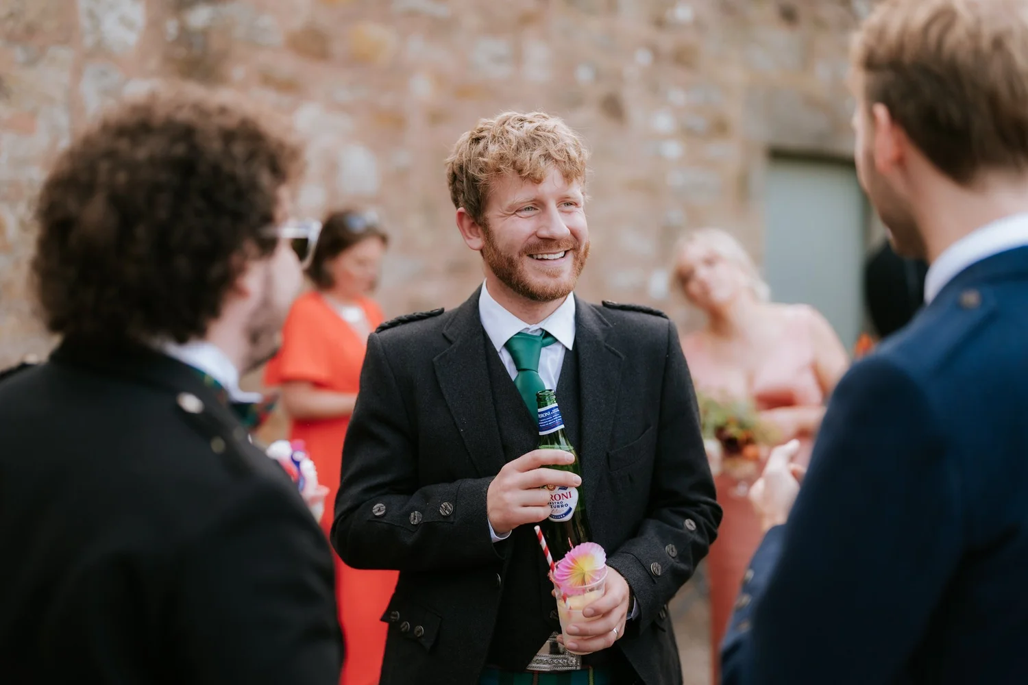 A guest in a dark waistcoat laughs with others while holding a drink in the courtyard.