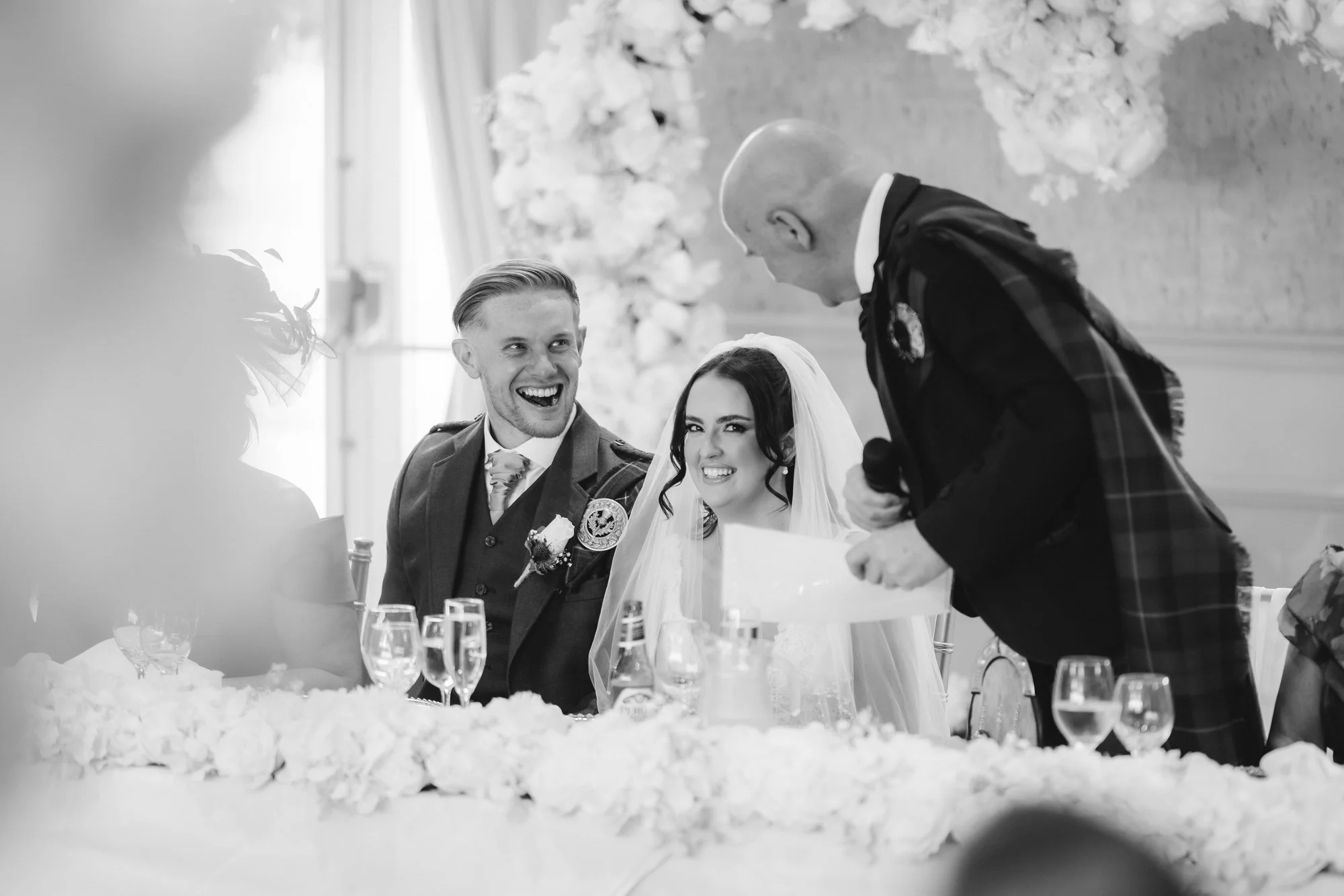 Kaitlyn and Scott laughing with a speaker at the top table, in black and white at Glenbervie House.