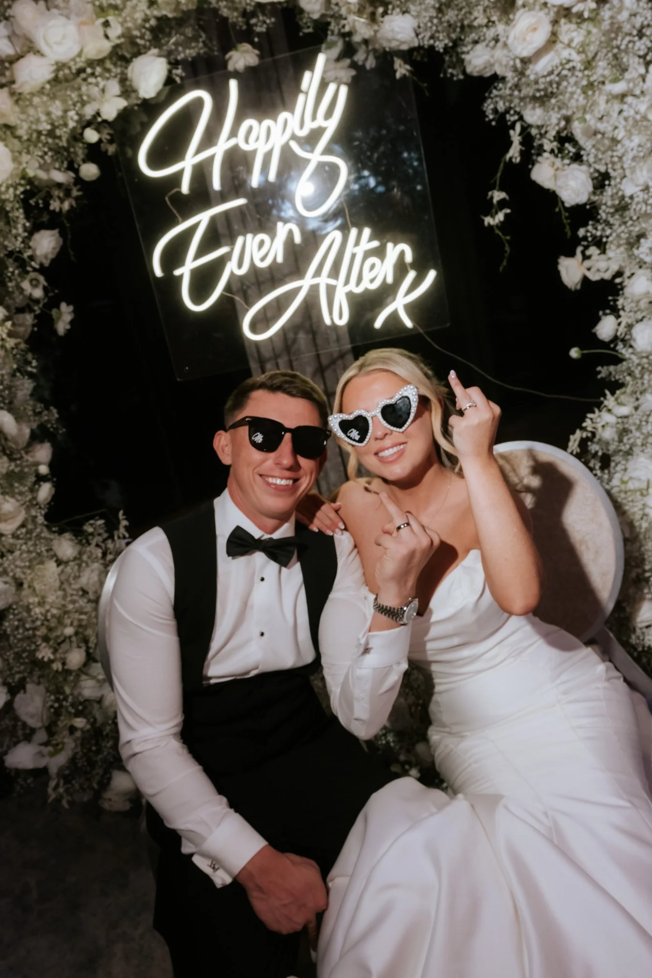 Aiden and Taylor wearing sunglasses by the wedding neon sign.