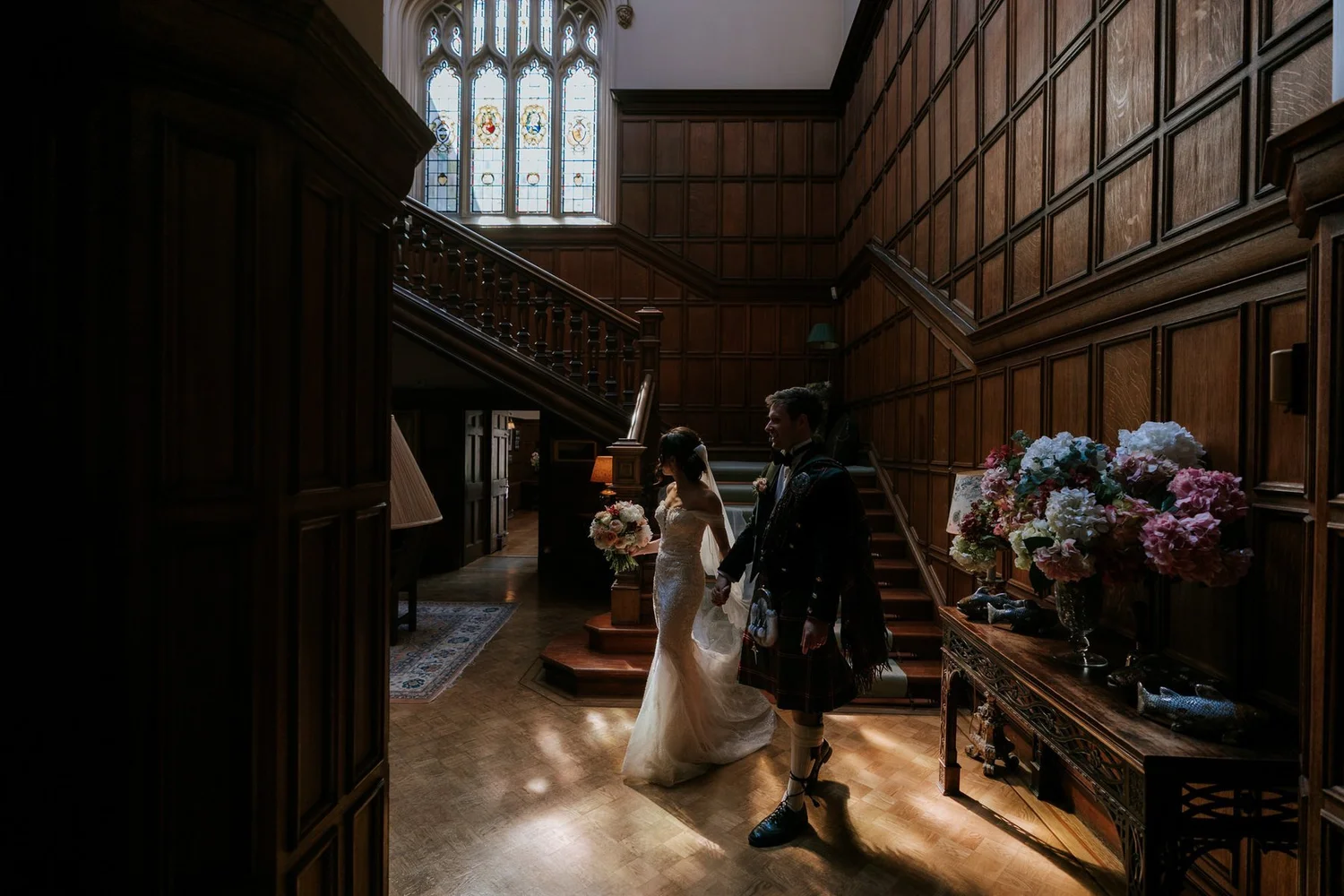 Kim and Kenneth walking through the wood-panelled hall beneath the stained-glass window at Dundas Castle.