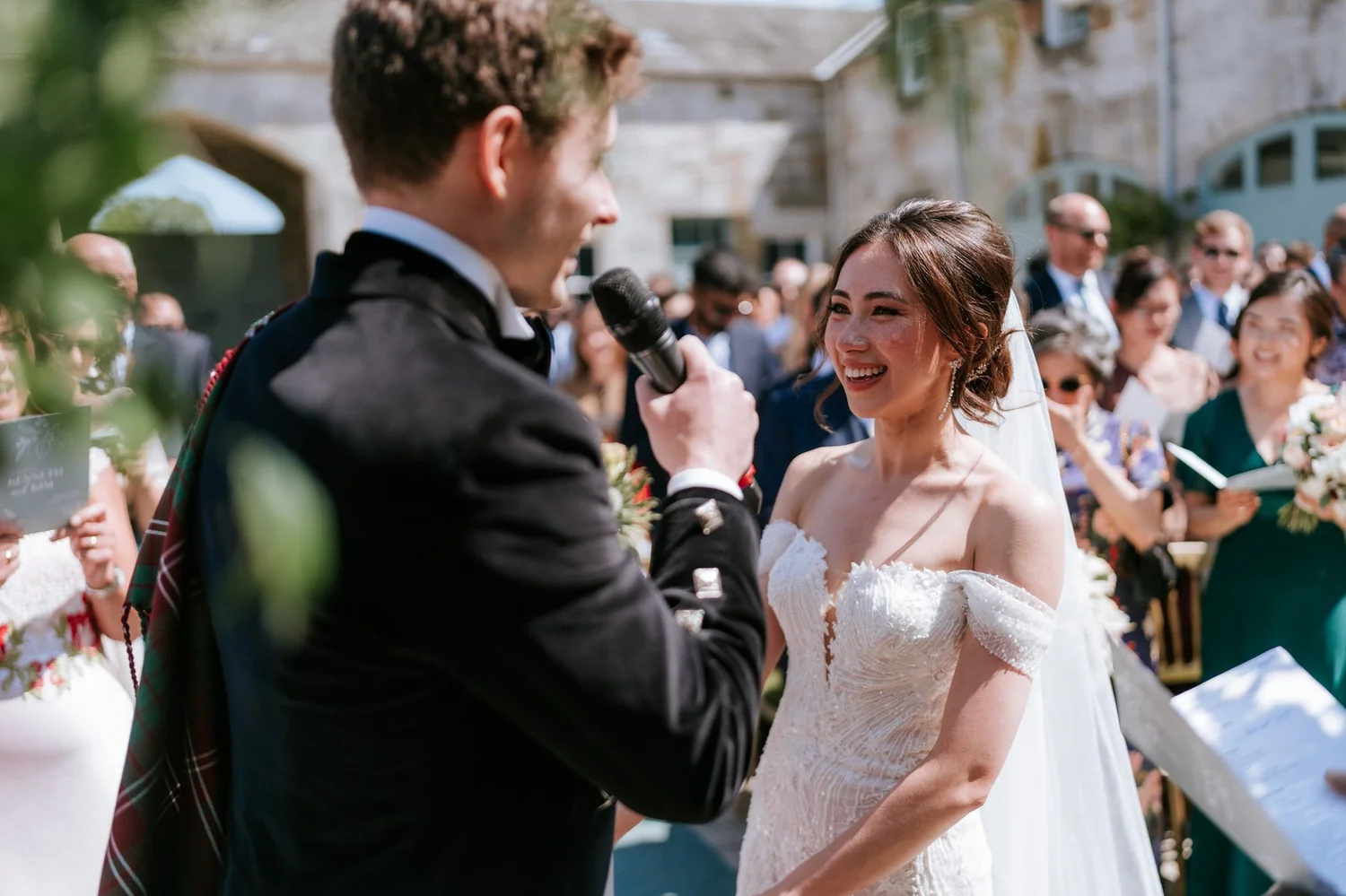 Kim smiling at Kenneth as he speaks into a microphone during their outdoor wedding ceremony.