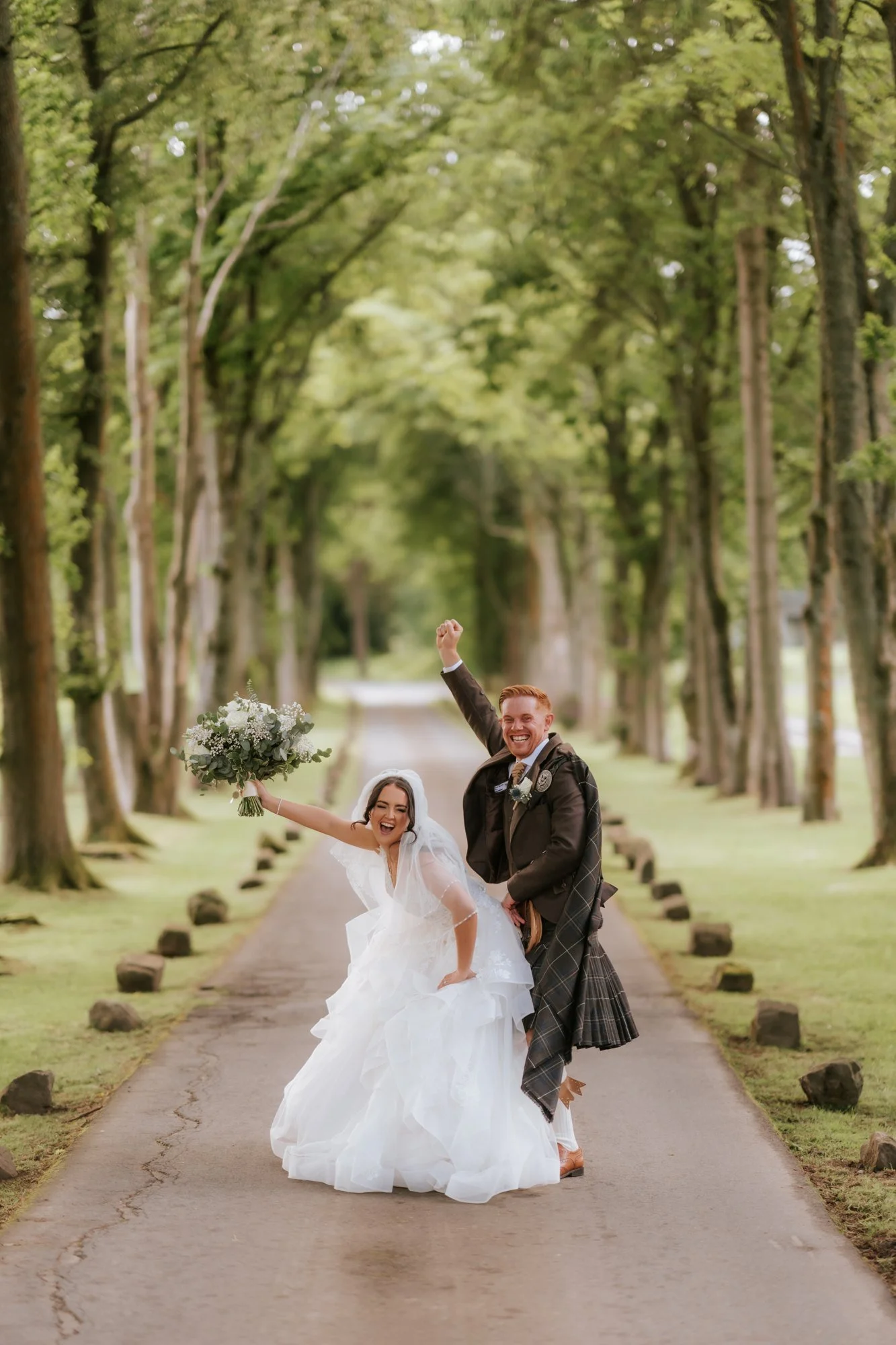 Kaitlyn raising her bouquet and Scott cheering on the tree-lined driveway at Glenbervie House.
