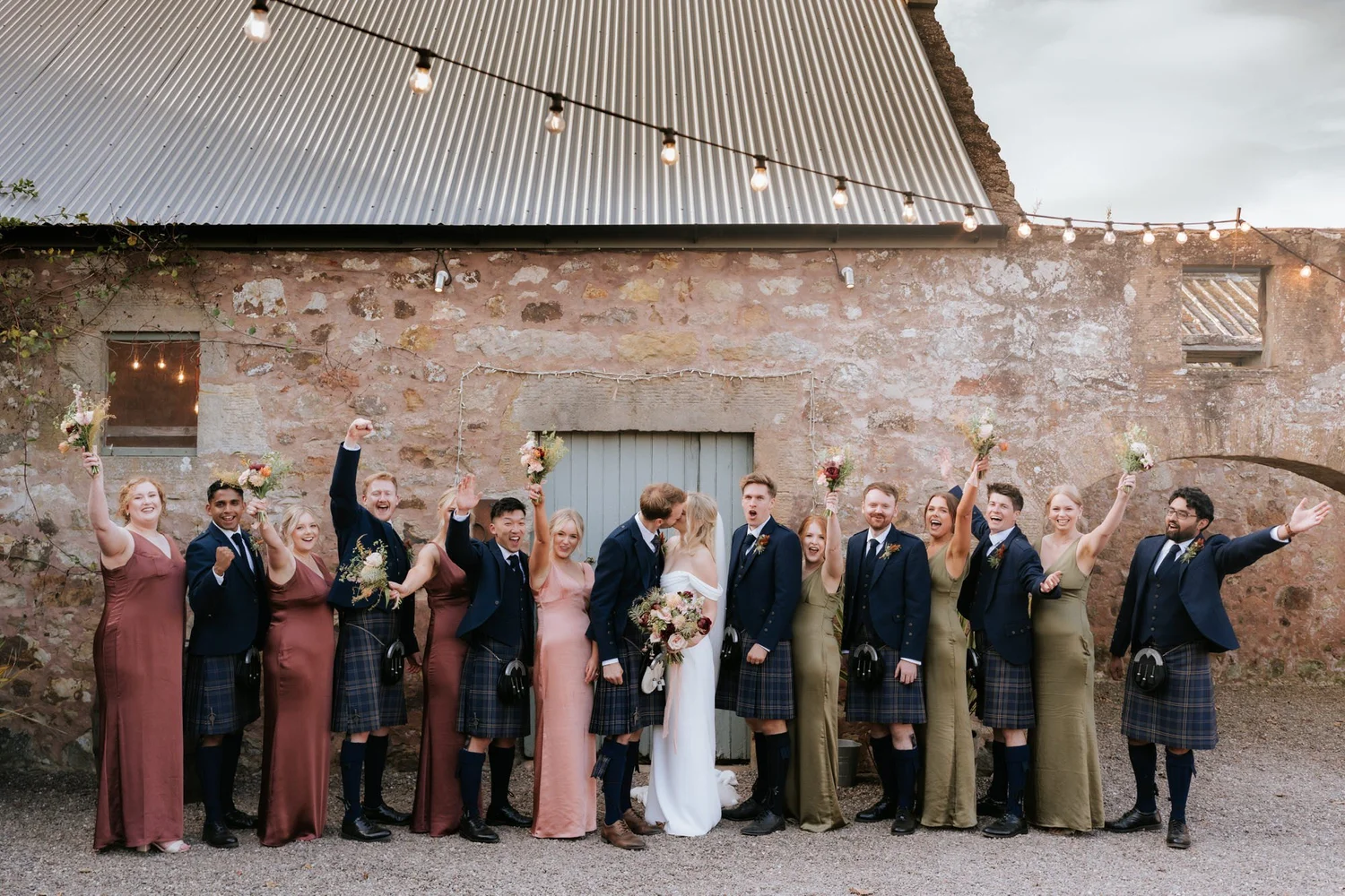 The wedding party cheer and raise their arms beside the couple in the courtyard.