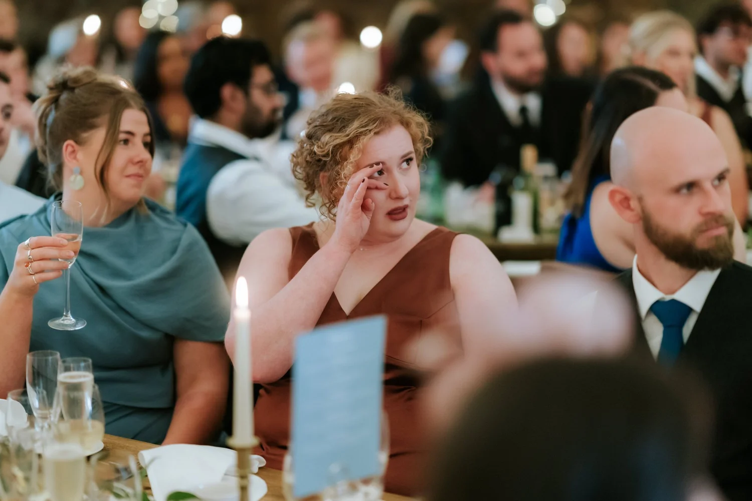 A guest in a burgundy dress touches the corner of her eye during the speeches.