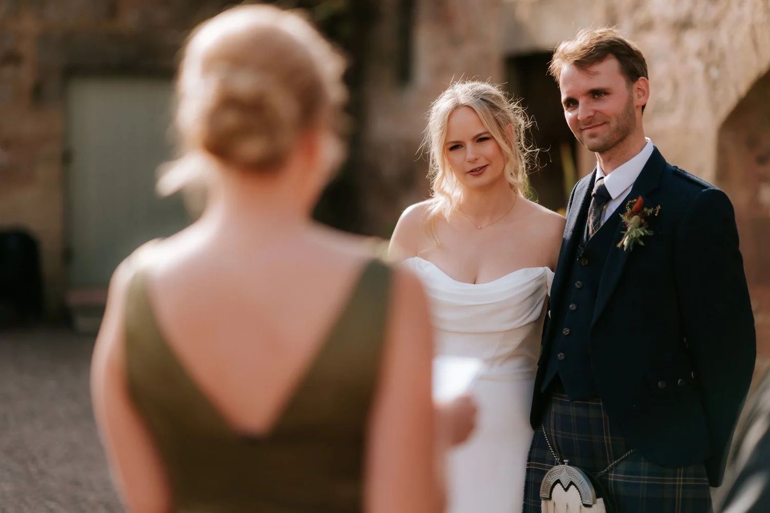 The bride smiles towards a reader during the outdoor ceremony, with the groom standing beside her.