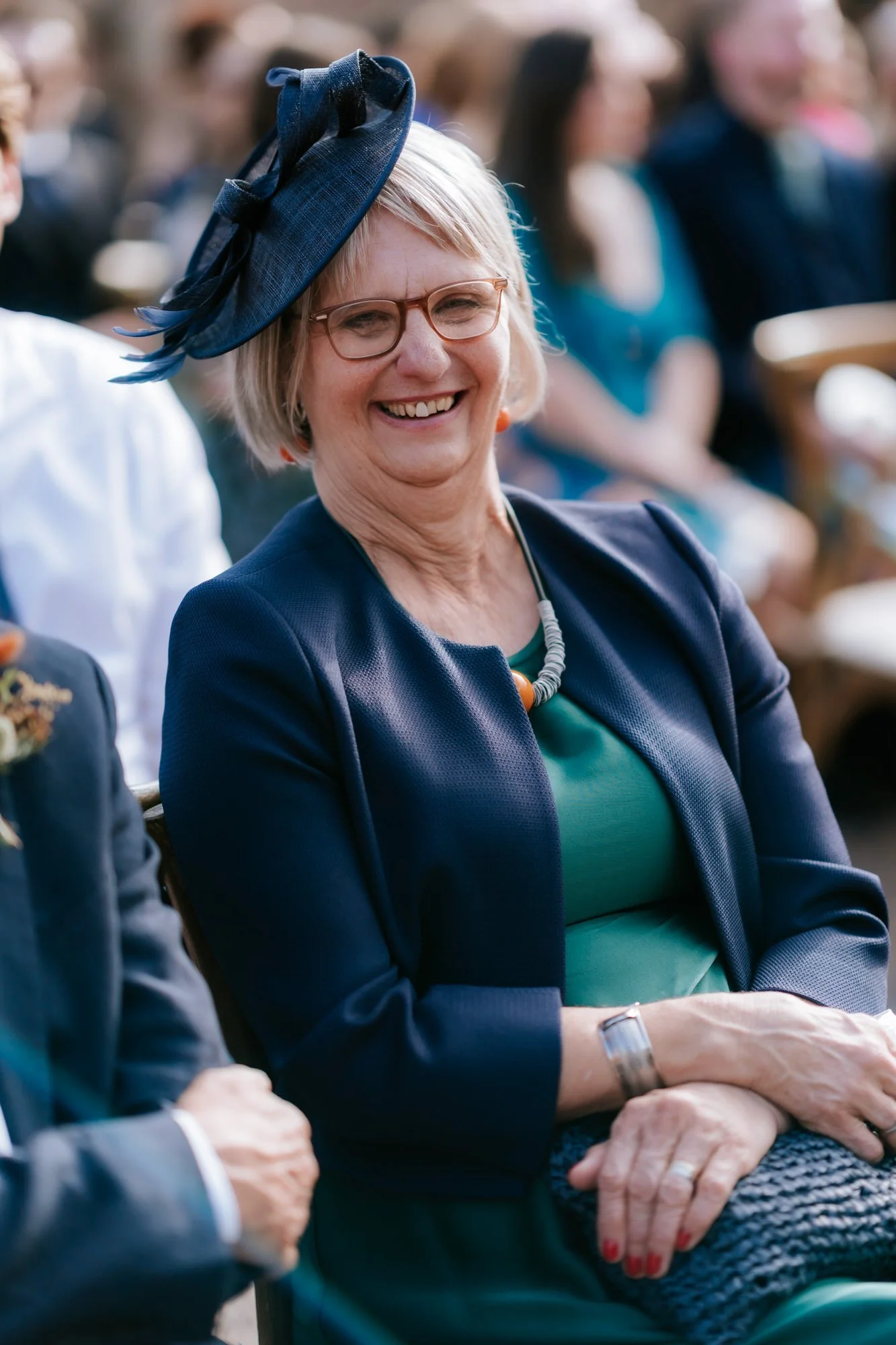 A guest in a teal dress and navy hat laughs while watching the outdoor wedding ceremony.