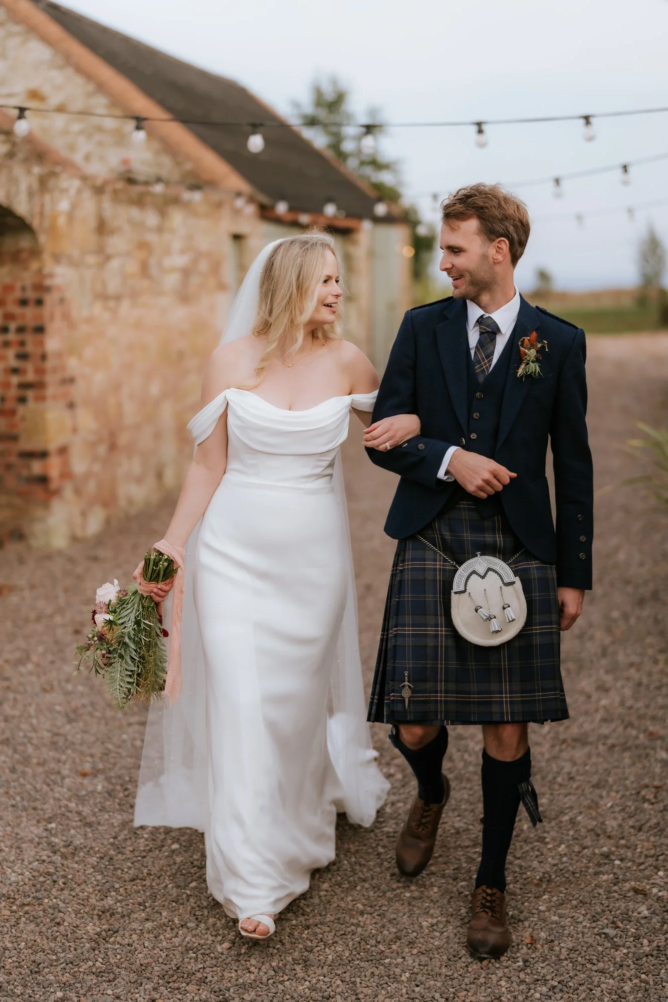 The couple walk arm in arm through the stone courtyard, the bride carrying her bouquet.