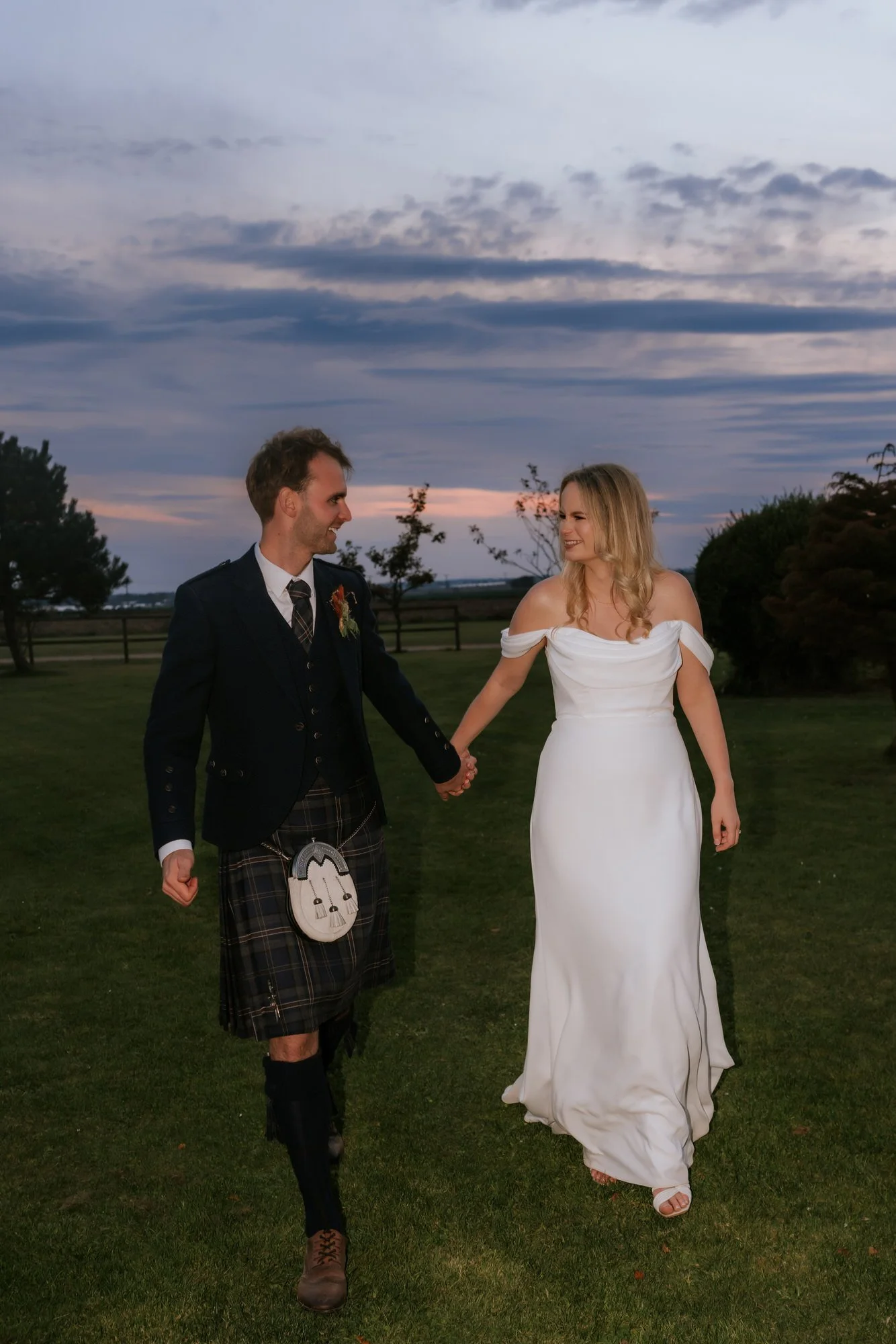 The couple smile at each other as they walk across the lawn beneath a blue evening sky.