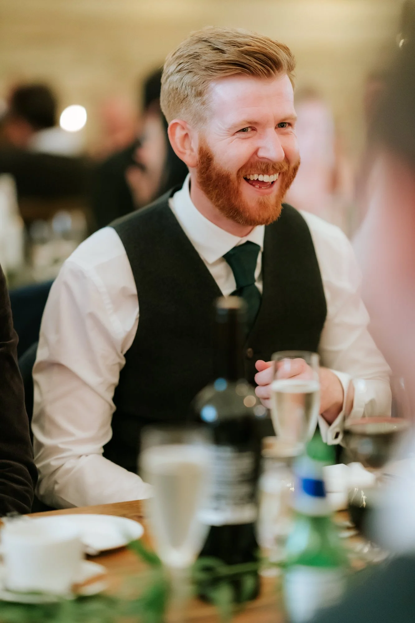 A bearded guest in a dark waistcoat and green tie laughs during the wedding speeches.