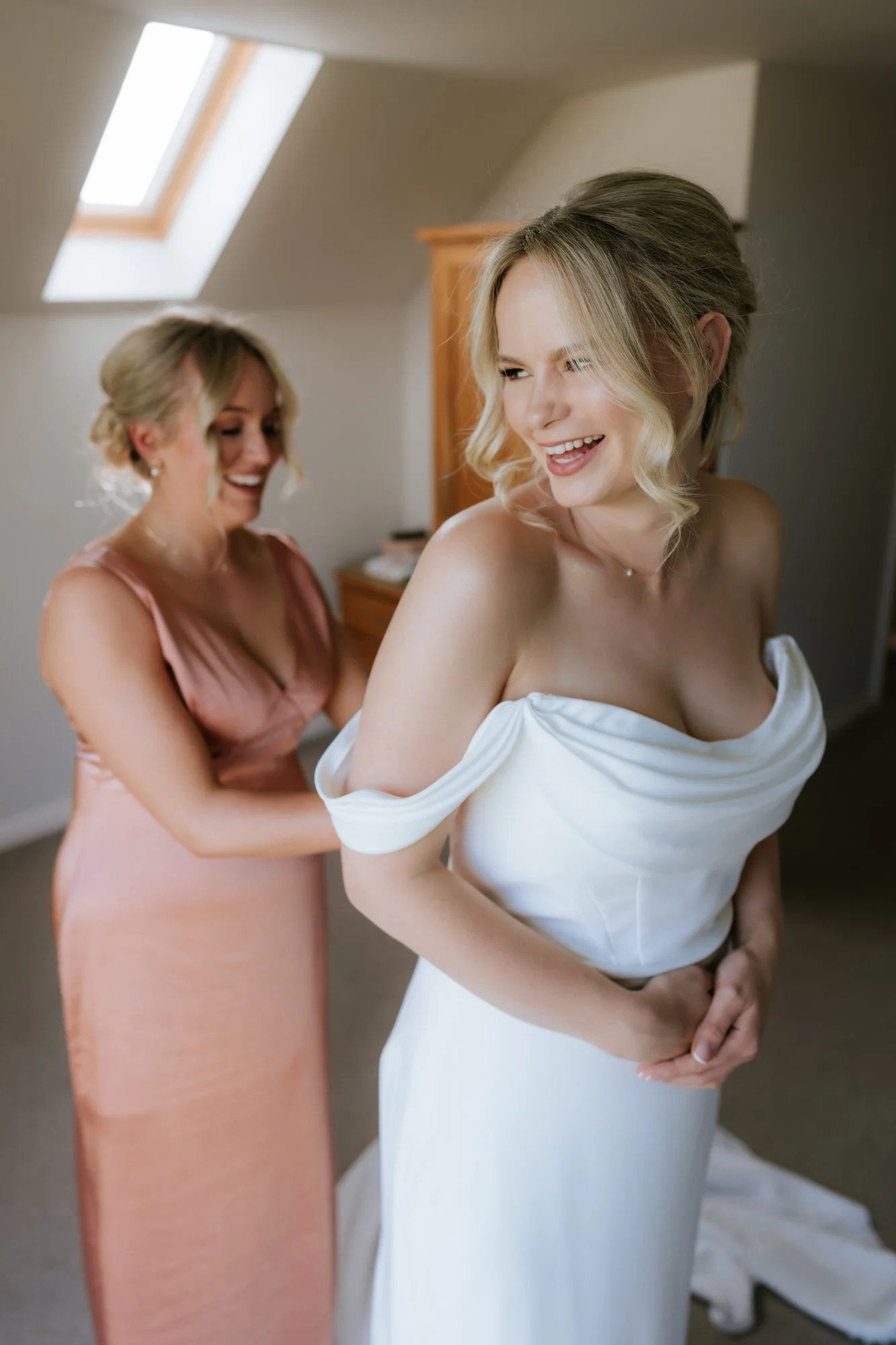 The bride smiles beside a window as a bridesmaid in a pink dress helps with her wedding dress.