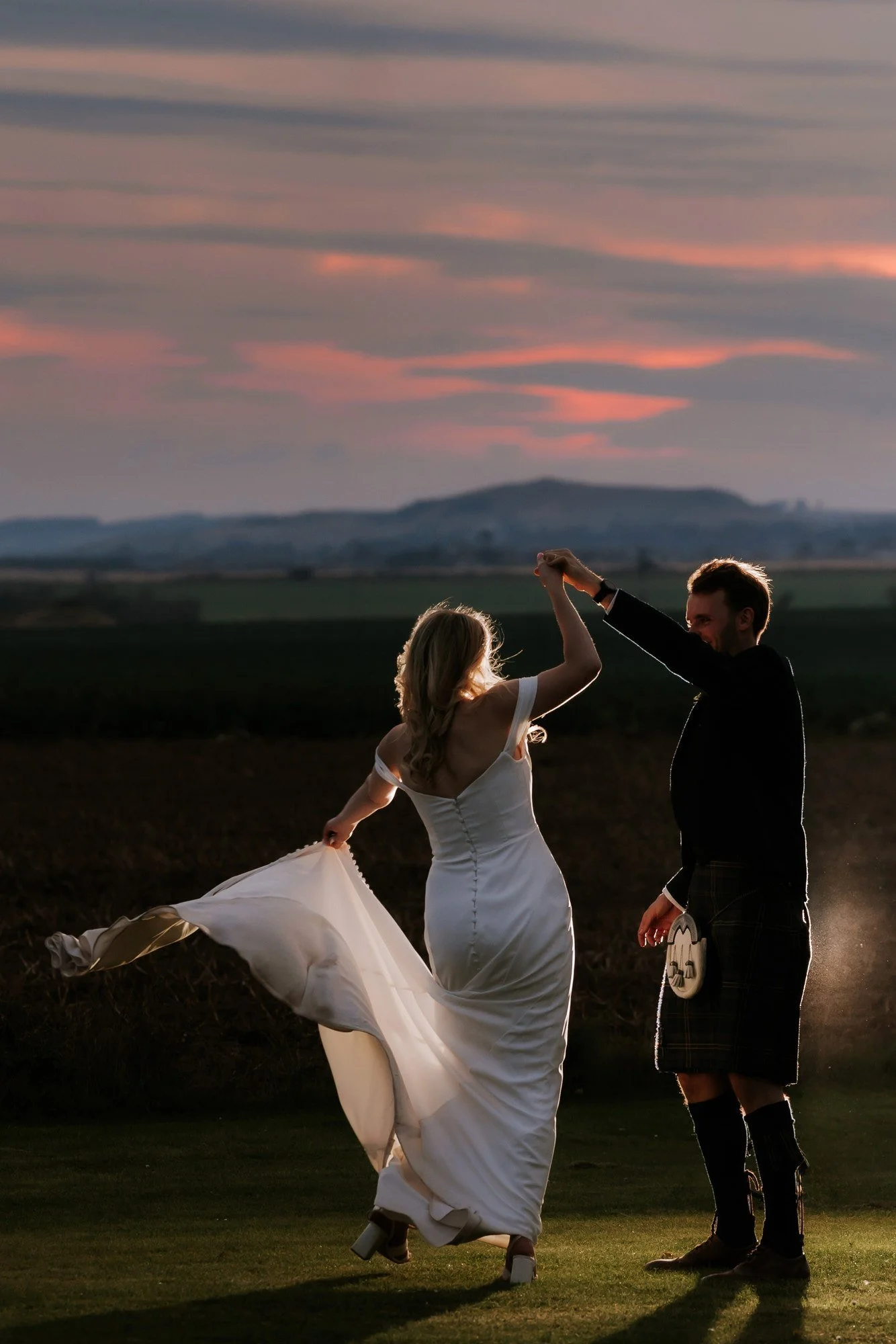 The groom raises the bride’s hand as she turns on the lawn beneath pink sunset clouds.
