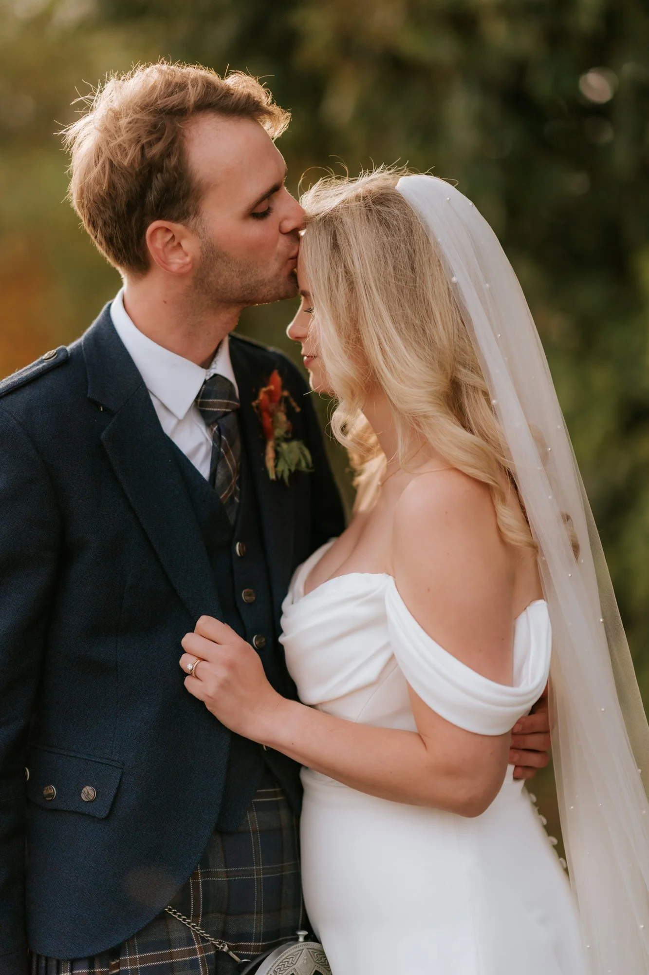 A close portrait of the groom kissing the bride’s forehead as she holds his jacket.