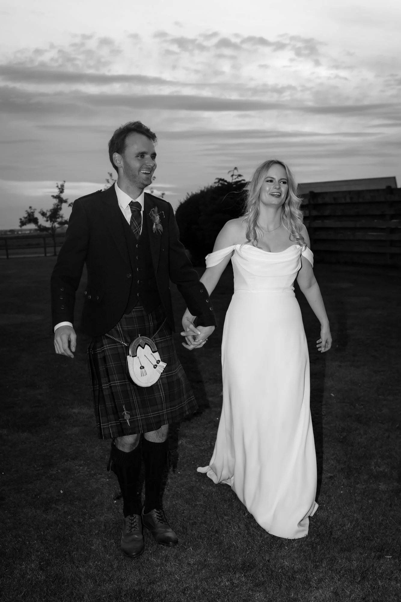 Black-and-white portrait of the couple walking hand in hand across the grass at dusk.