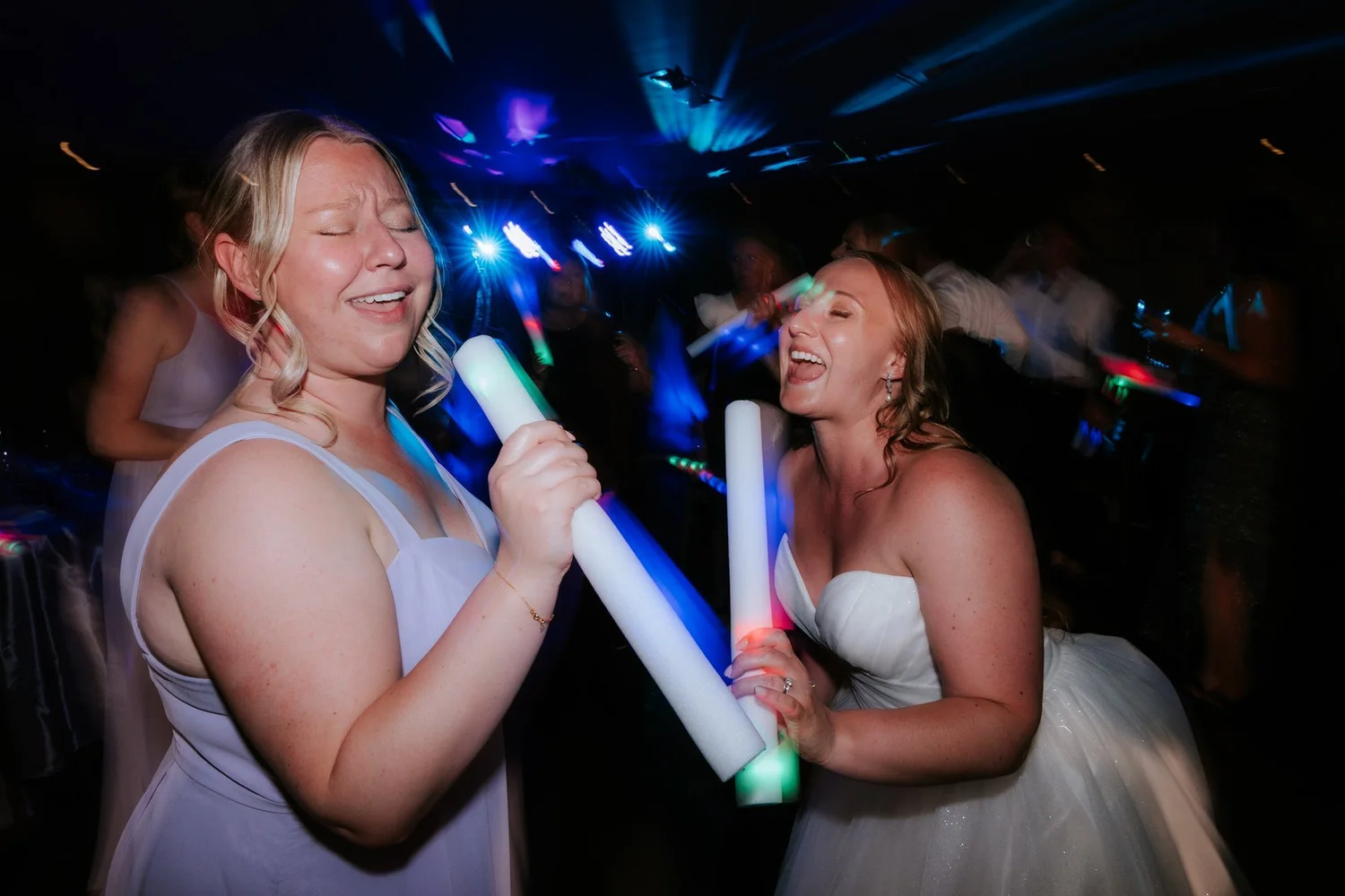 Allie and a bridesmaid singing along and laughing with glowing party sticks on the dance floor.