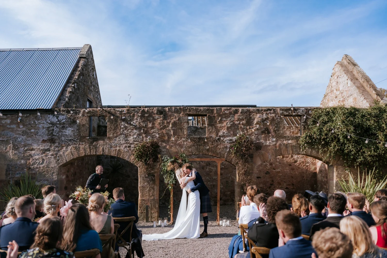 The couple share a kiss in front of the stone arches as guests watch the ceremony.