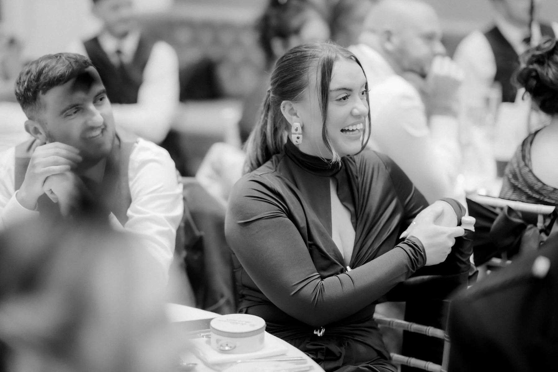 Wedding guests laughing and applauding around a reception table, photographed in black and white.