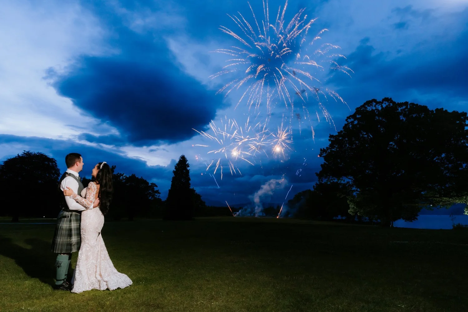 Cristina and Connor kissing on the lawn as fireworks burst above Cameron House at dusk.
