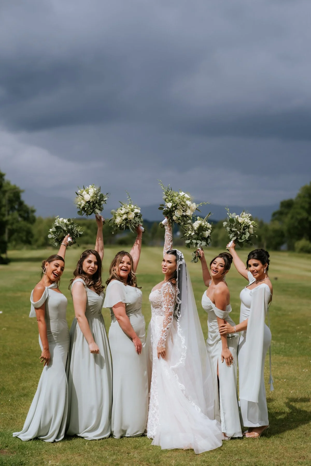 Cristina and her bridesmaids raising their bouquets on the lawn at Cameron House.