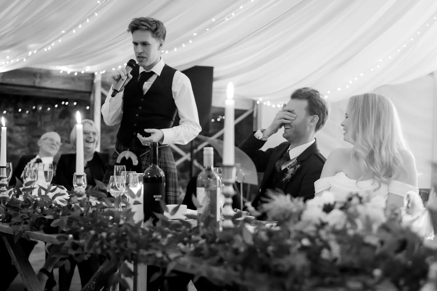 Black-and-white photograph of a wedding speech, with the groom covering his face and the bride looking on.