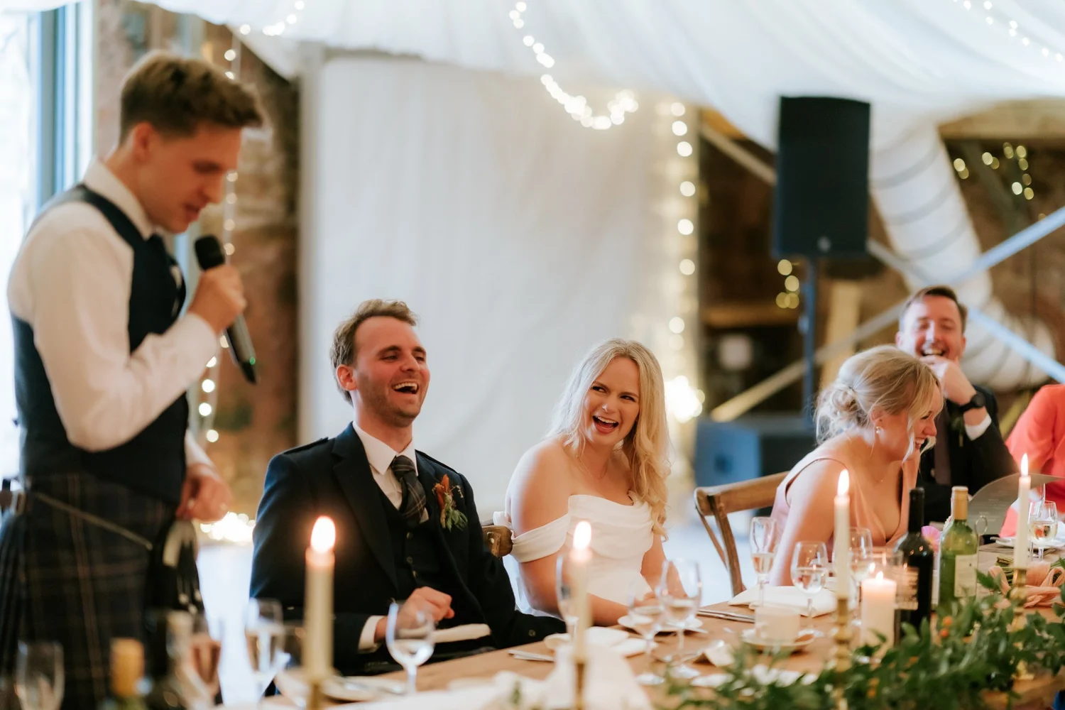 A speaker holds a microphone as the couple and guests laugh at the candlelit wedding table.