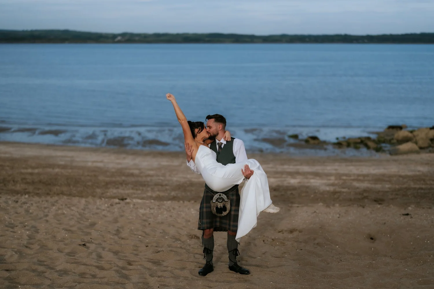 Dougie carrying Lois as she raises her arm and kisses him beside the sea.