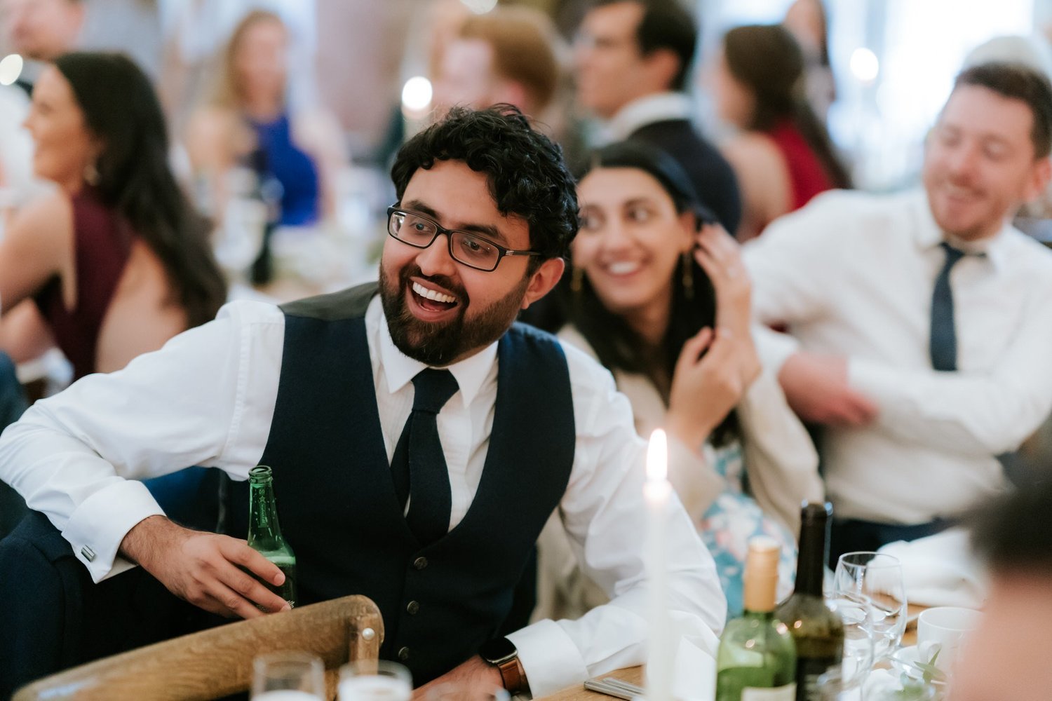 A guest wearing glasses and a dark waistcoat laughs while holding a green bottle at the reception table.