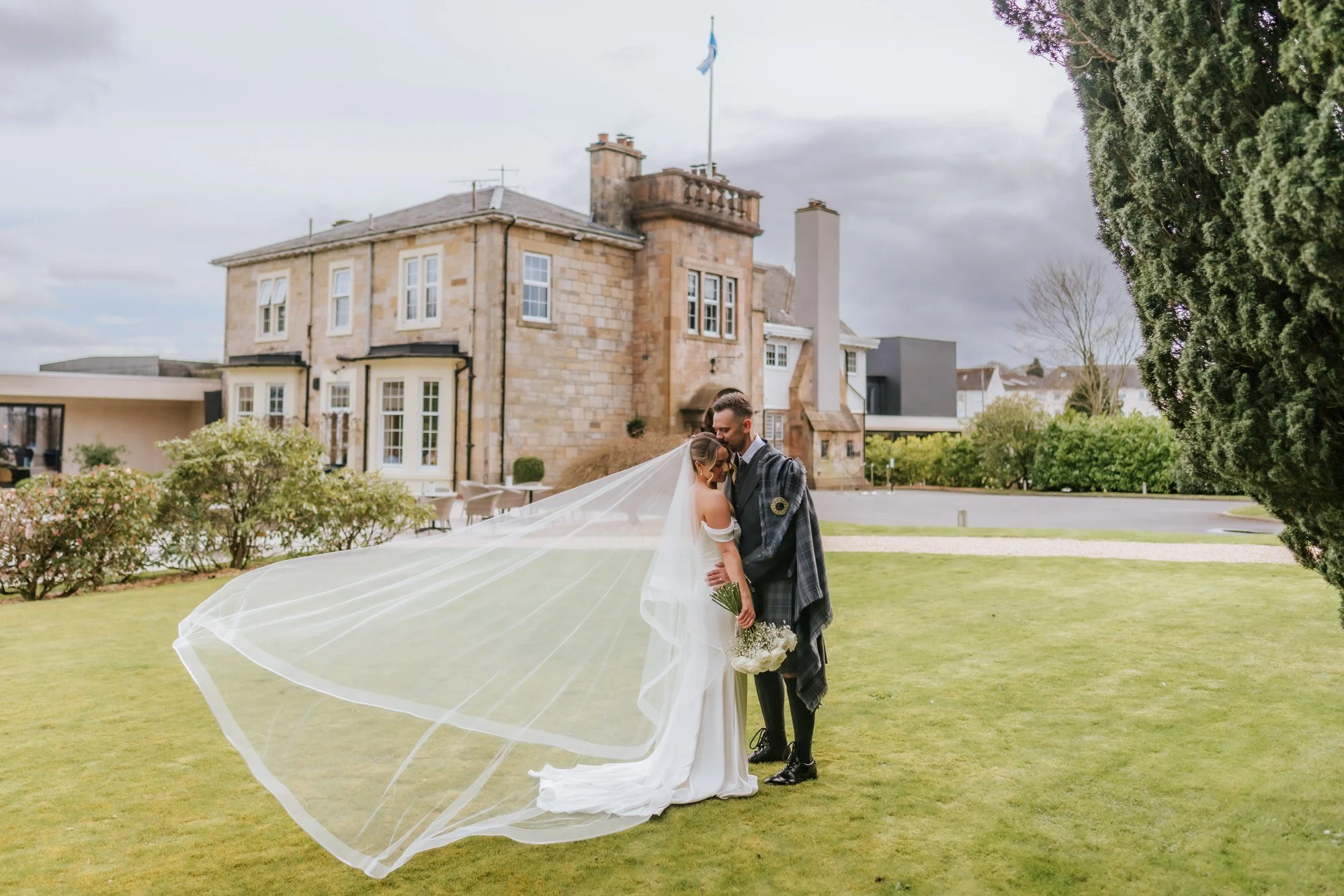 Elle and Adam beside Dalmeny Park House Hotel with her veil flowing behind them