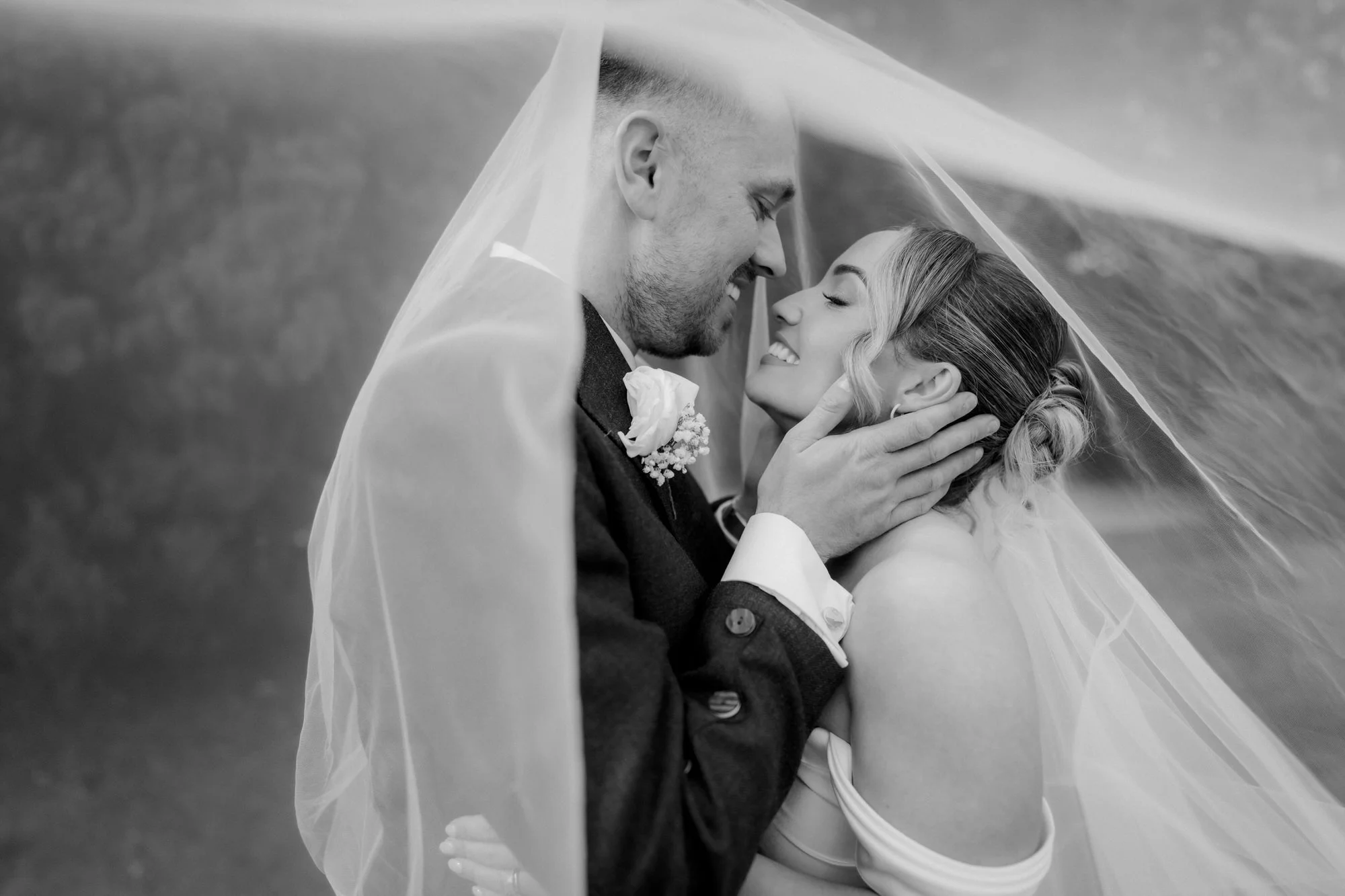 A close black-and-white portrait of Elle and Adam smiling beneath her veil