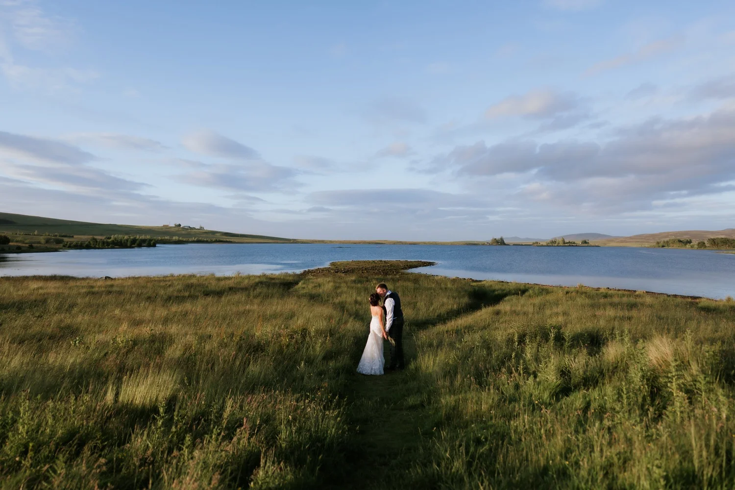Jamie and Lauren standing in long grass beside the reservoir at Cairns Farm Estate, with the Pentland Hills beyond.