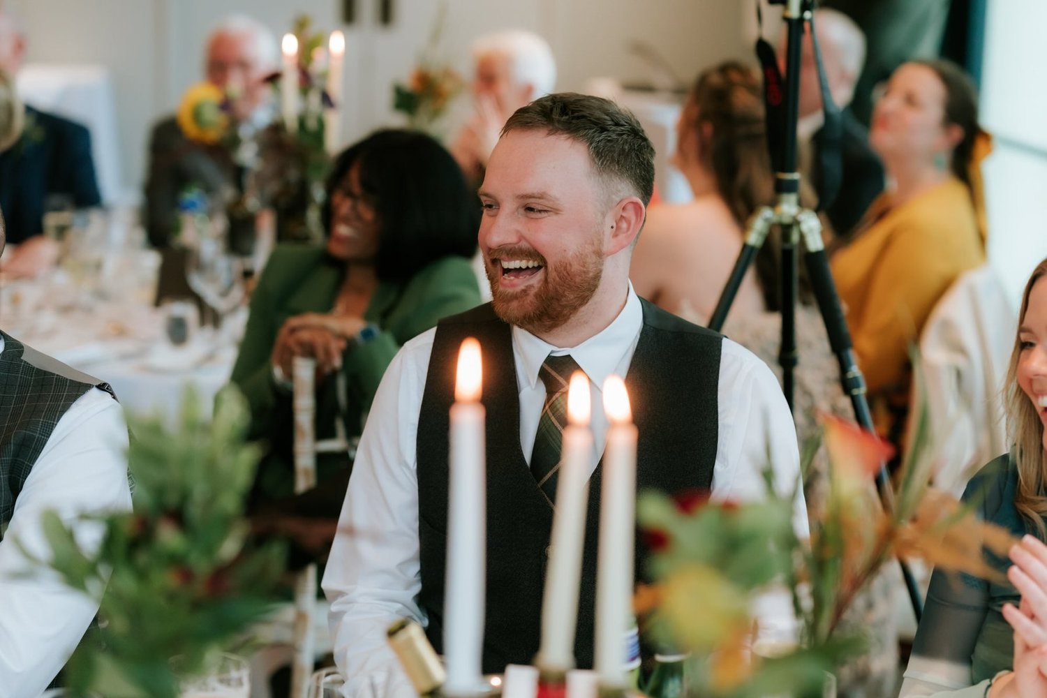 A bearded guest laughing across the candlelit wedding breakfast table.