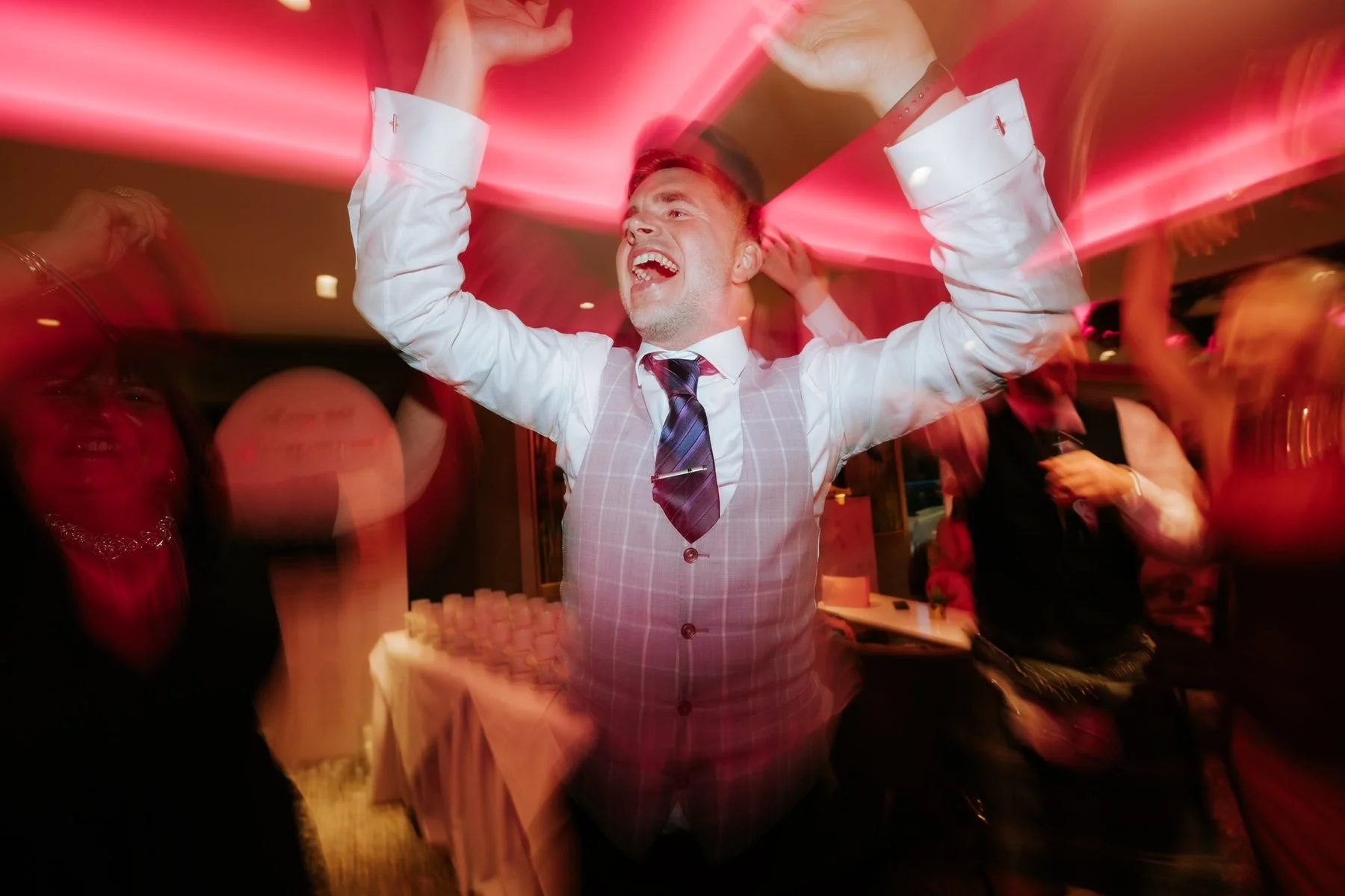 A guest dancing with both arms raised beneath the pink reception lighting.