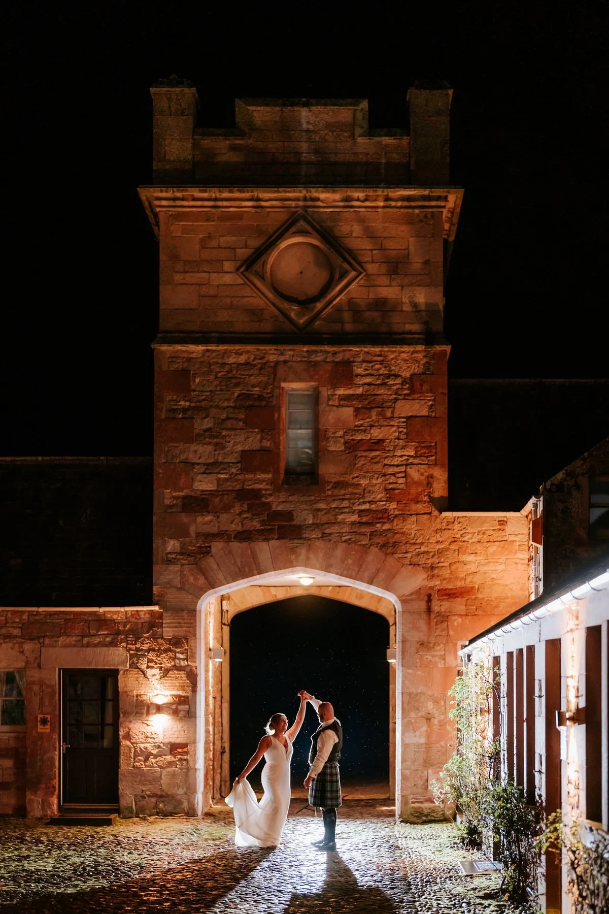 Kendall and Ewen beneath an illuminated stone archway in the hotel grounds after dark.