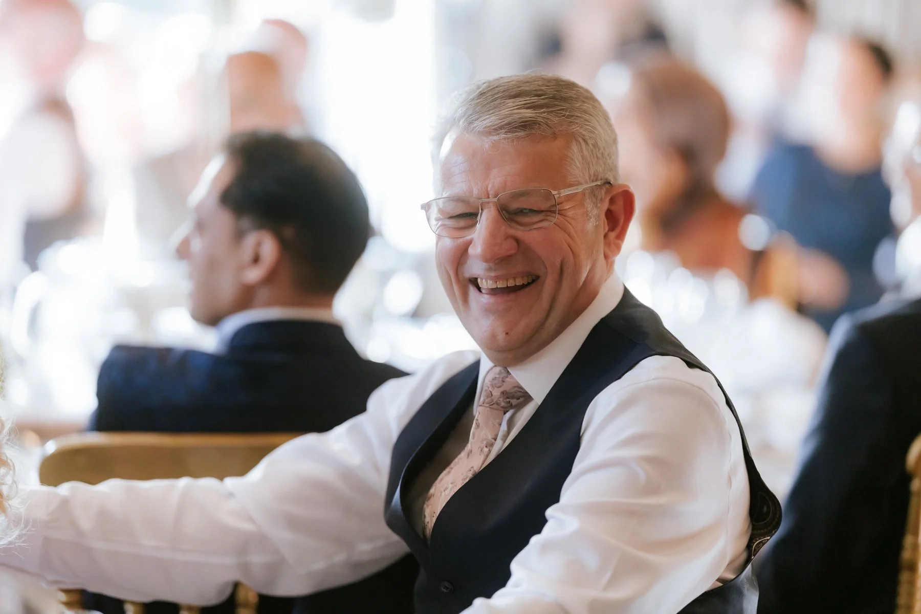 A guest in a white shirt and dark waistcoat laughing during the speeches.