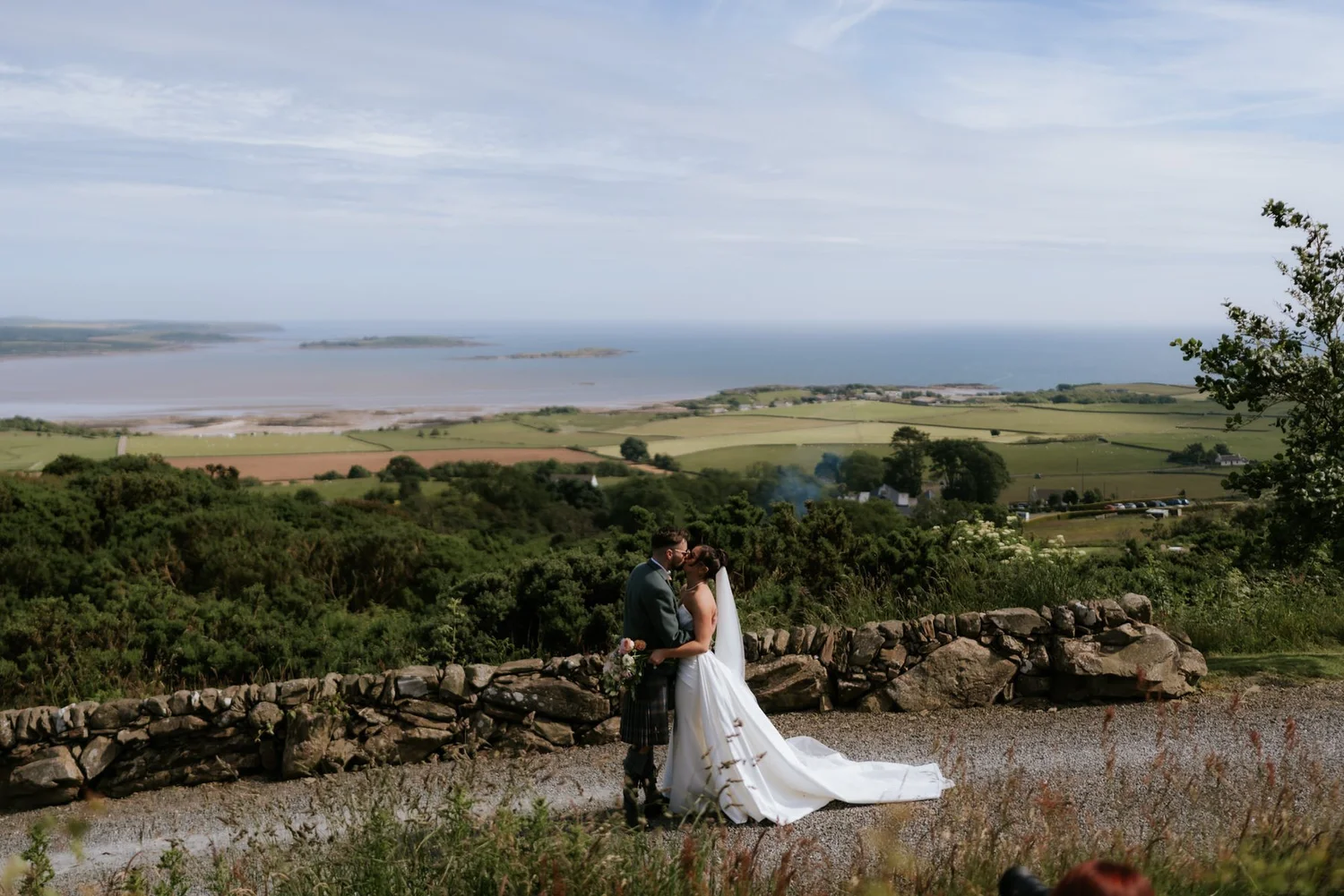 Lois and Dougie embracing beside a stone wall with sweeping coastal views from GG’s Yard.