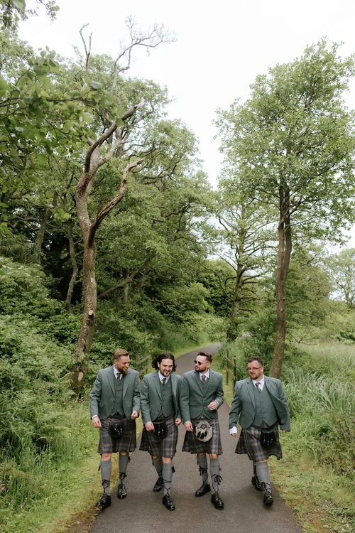 Dougie and his groomsmen walking together along a tree-lined path in their kilts.