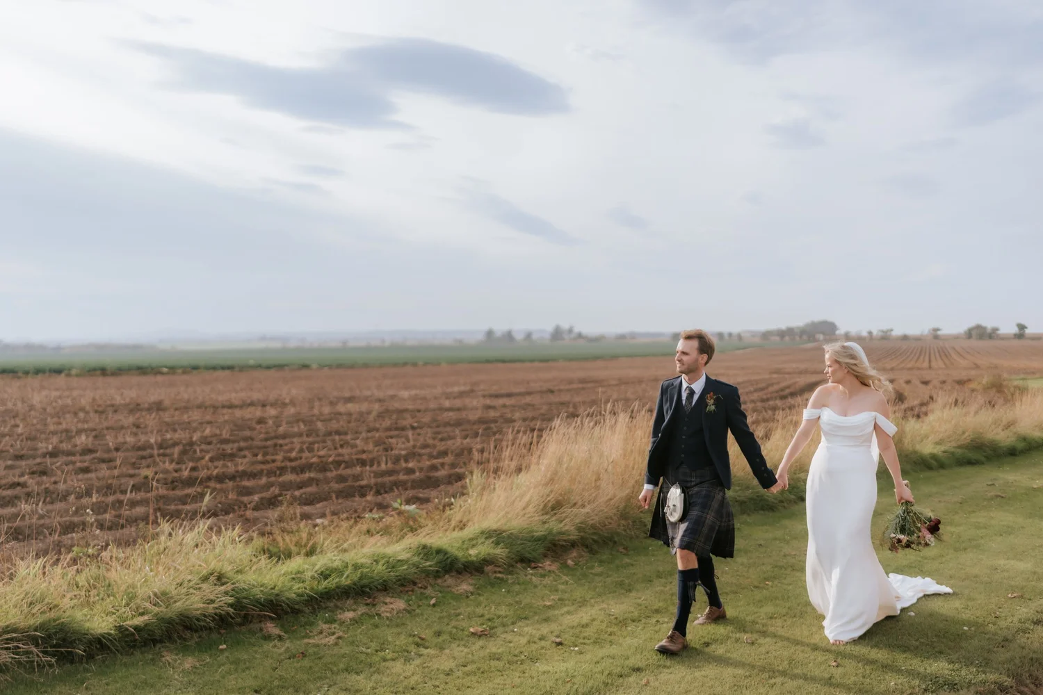The couple walk hand in hand along a grassy strip beside a ploughed field.