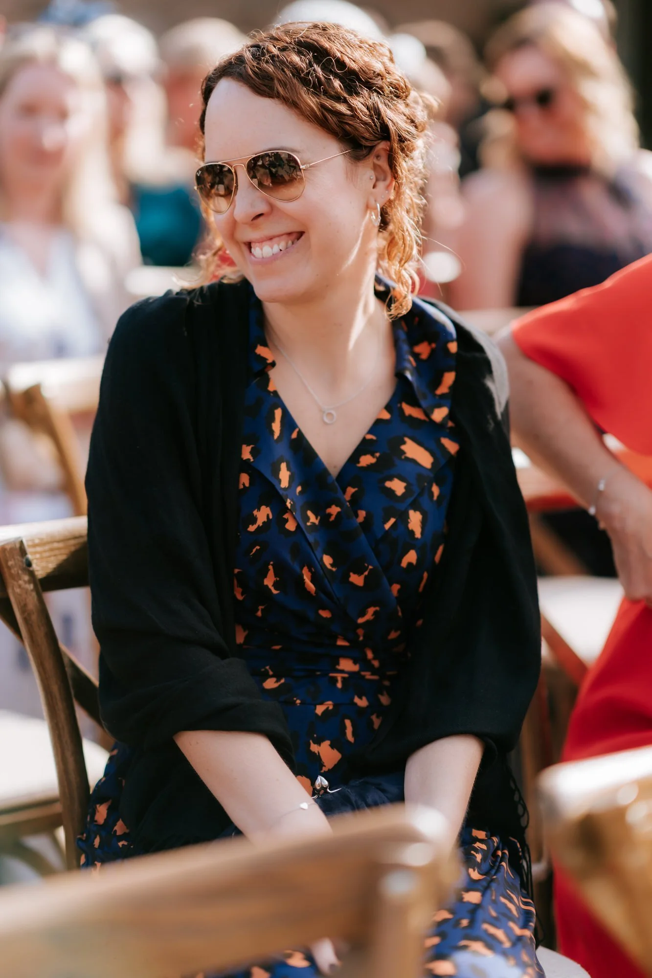 A guest in sunglasses, a floral dress and a dark jacket smiles during the outdoor ceremony.