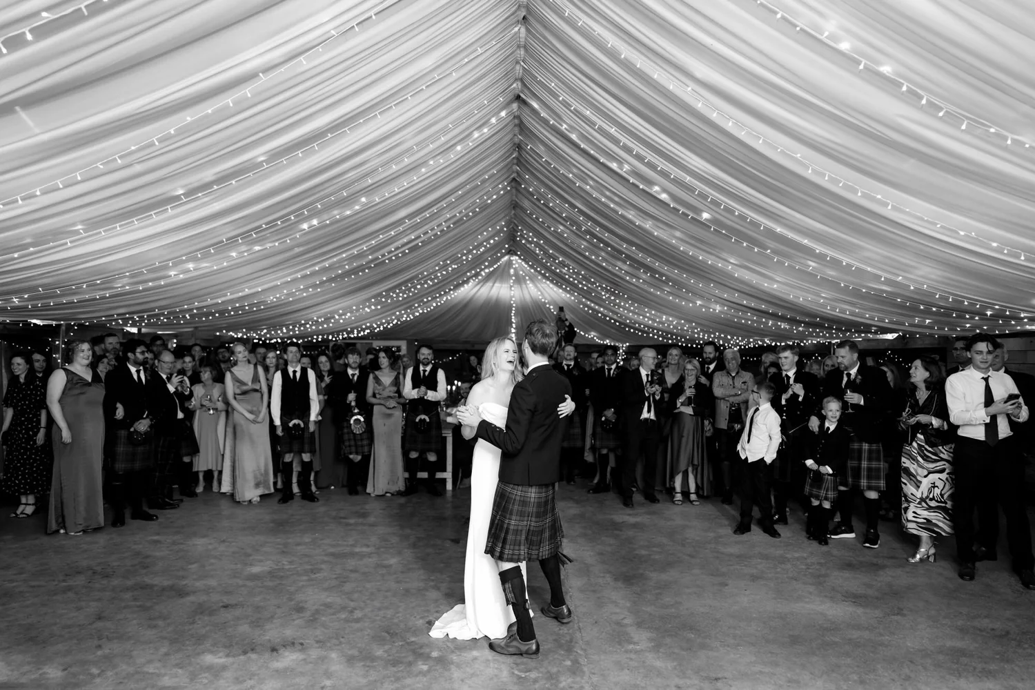 Black-and-white photograph of the couple dancing beneath the marquee canopy as guests watch around them.