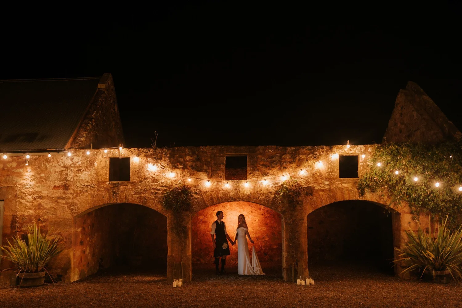 The couple stand beneath a warmly lit stone arch at The Cow Shed Crail, with festoon lights overhead.