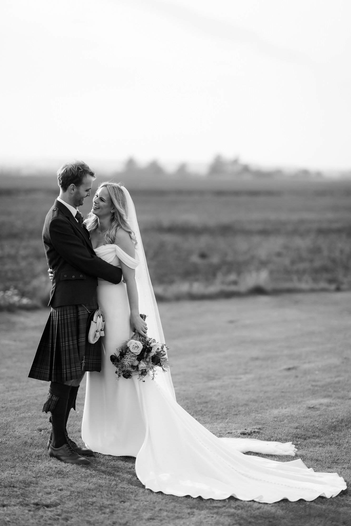Black-and-white full-length portrait of the couple embracing, with the bride’s train on the grass.