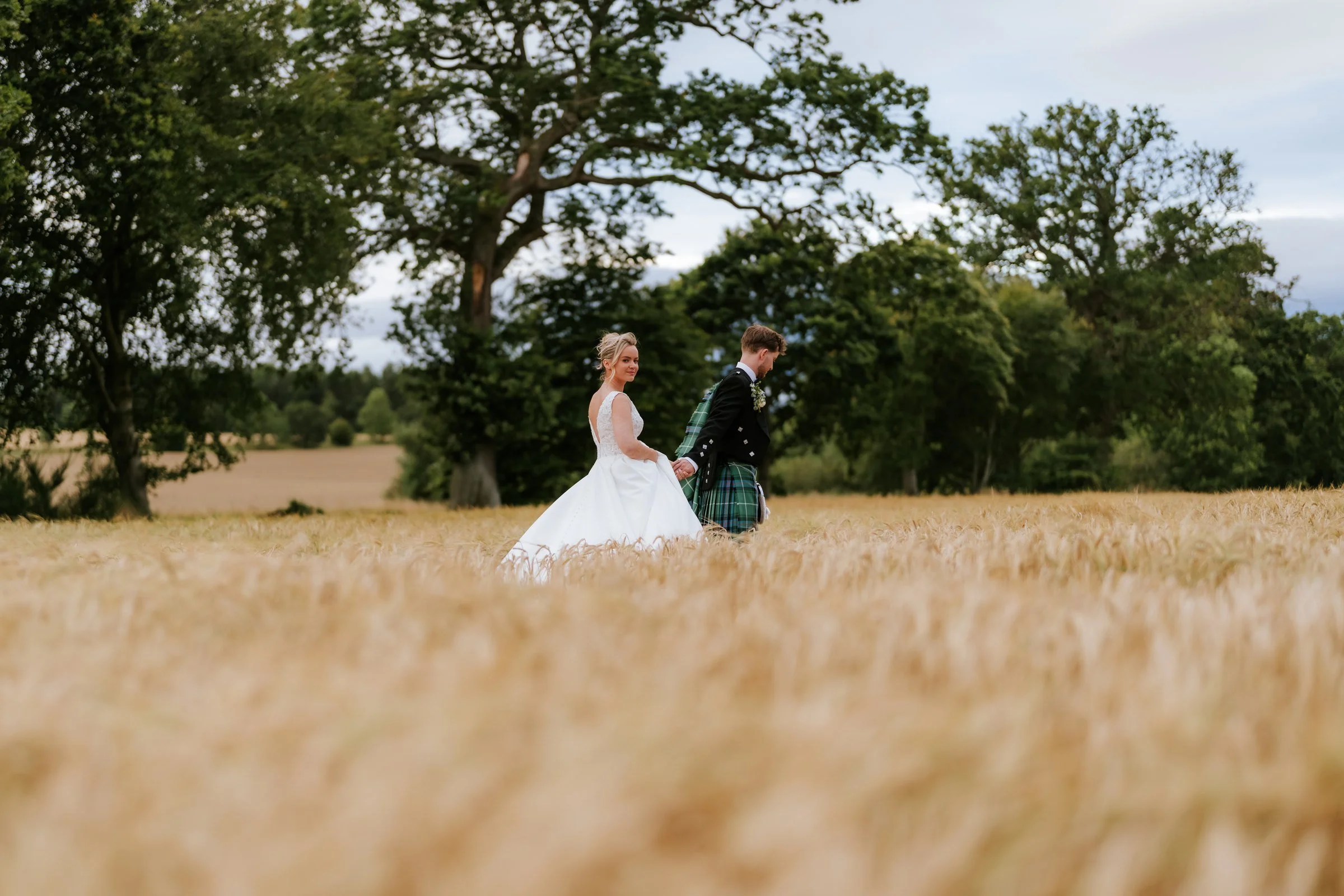 Lilian and Luke holding hands in a golden field beneath the trees at Rossie on the Earn.